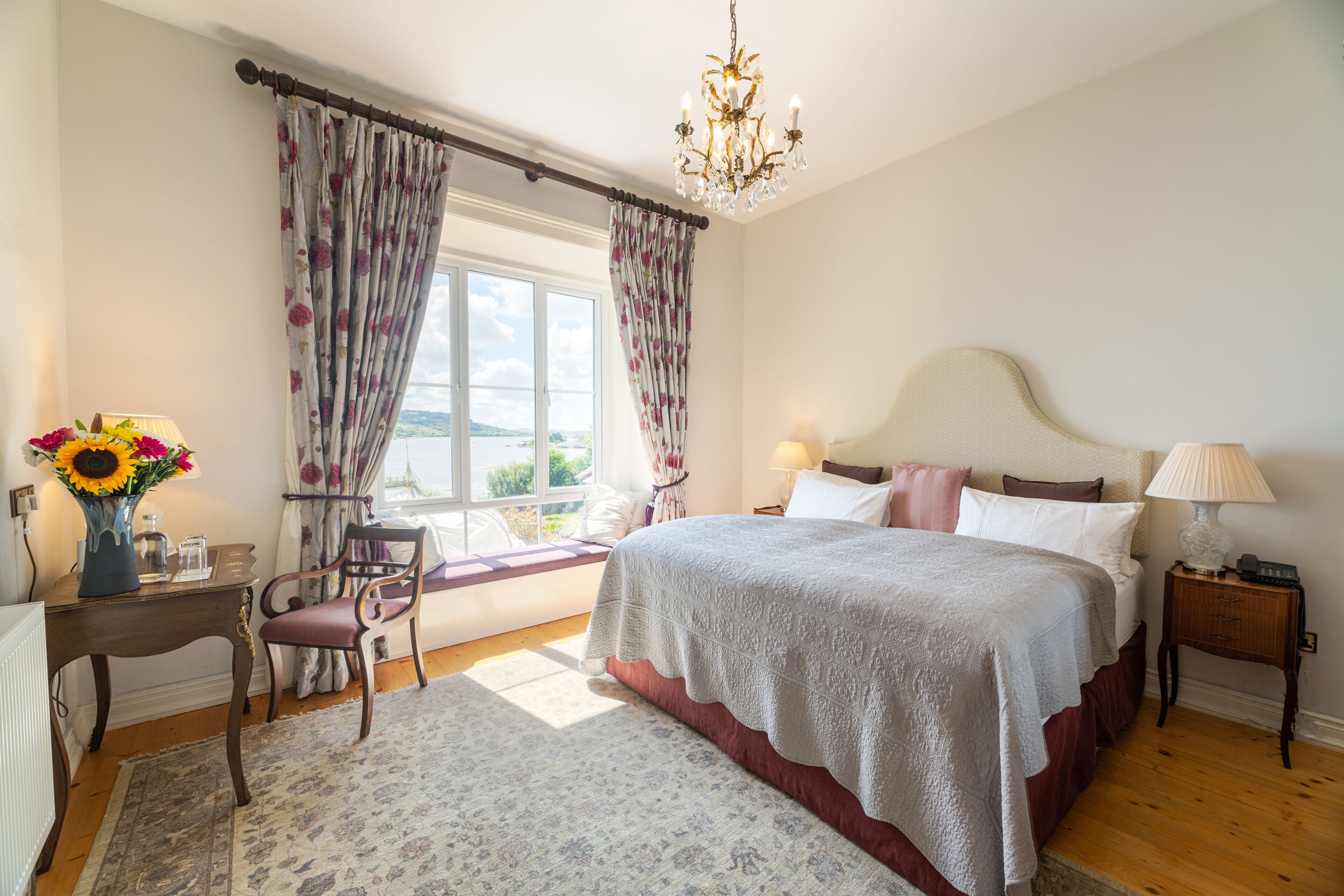 Un dormitorio con una cama grande, una mesita de noche, una silla, una ventana con cortinas y un candelabro.