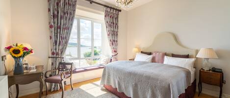 Un dormitorio con una cama grande, una mesita de noche, una silla, una ventana con cortinas y un candelabro.