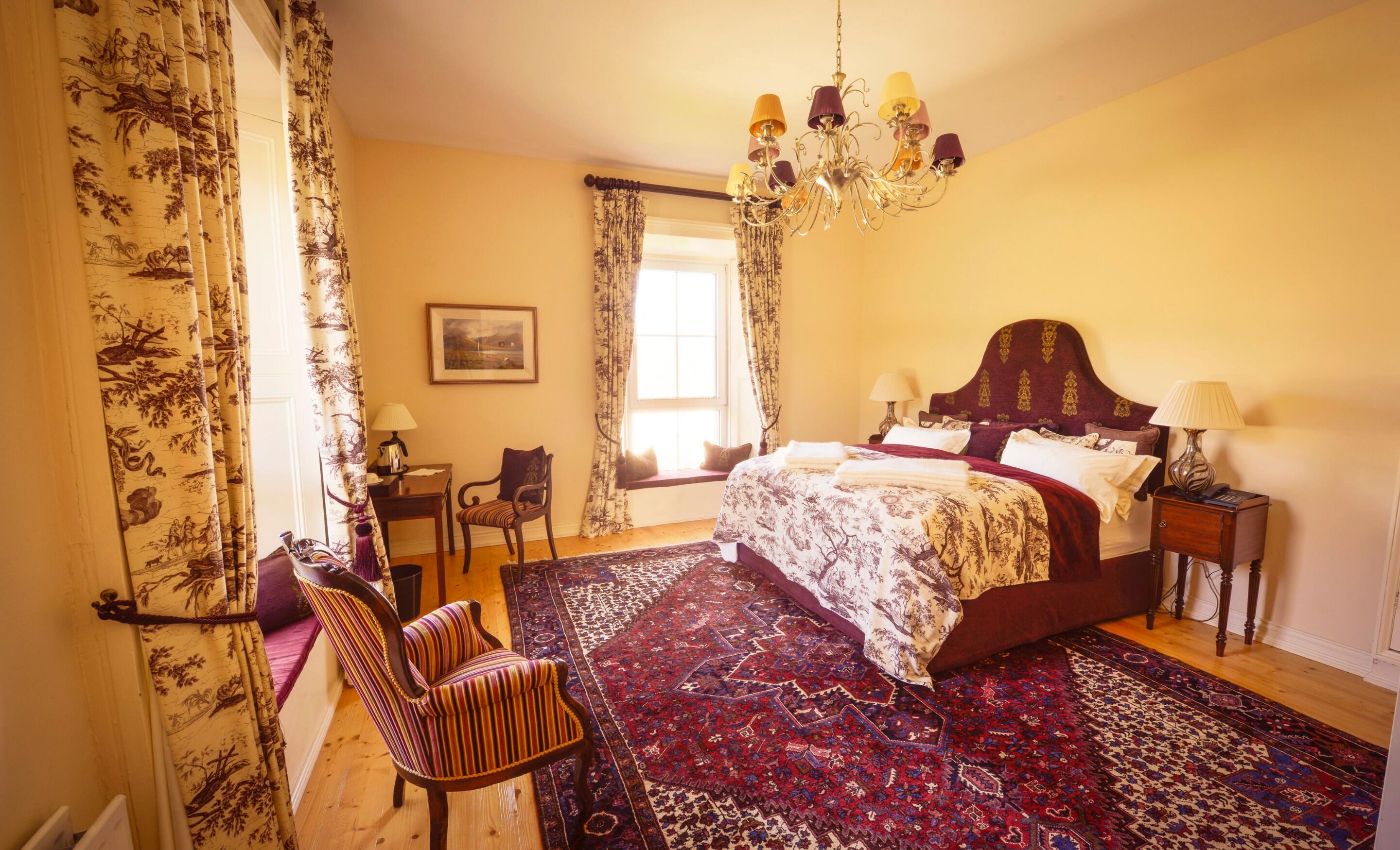 Un dormitorio con una cama grande, una araña, un escritorio con silla y una ventana con cortinas.