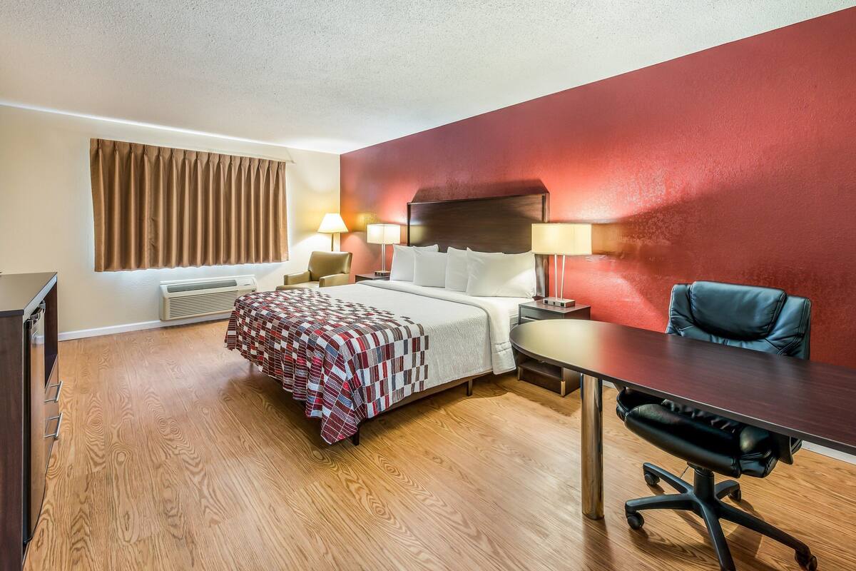 Ein Hotelzimmer mit einem Bett, einem Schreibtisch, einem Stuhl, einer Lampe und einem fensterbepflanzten Fenster.