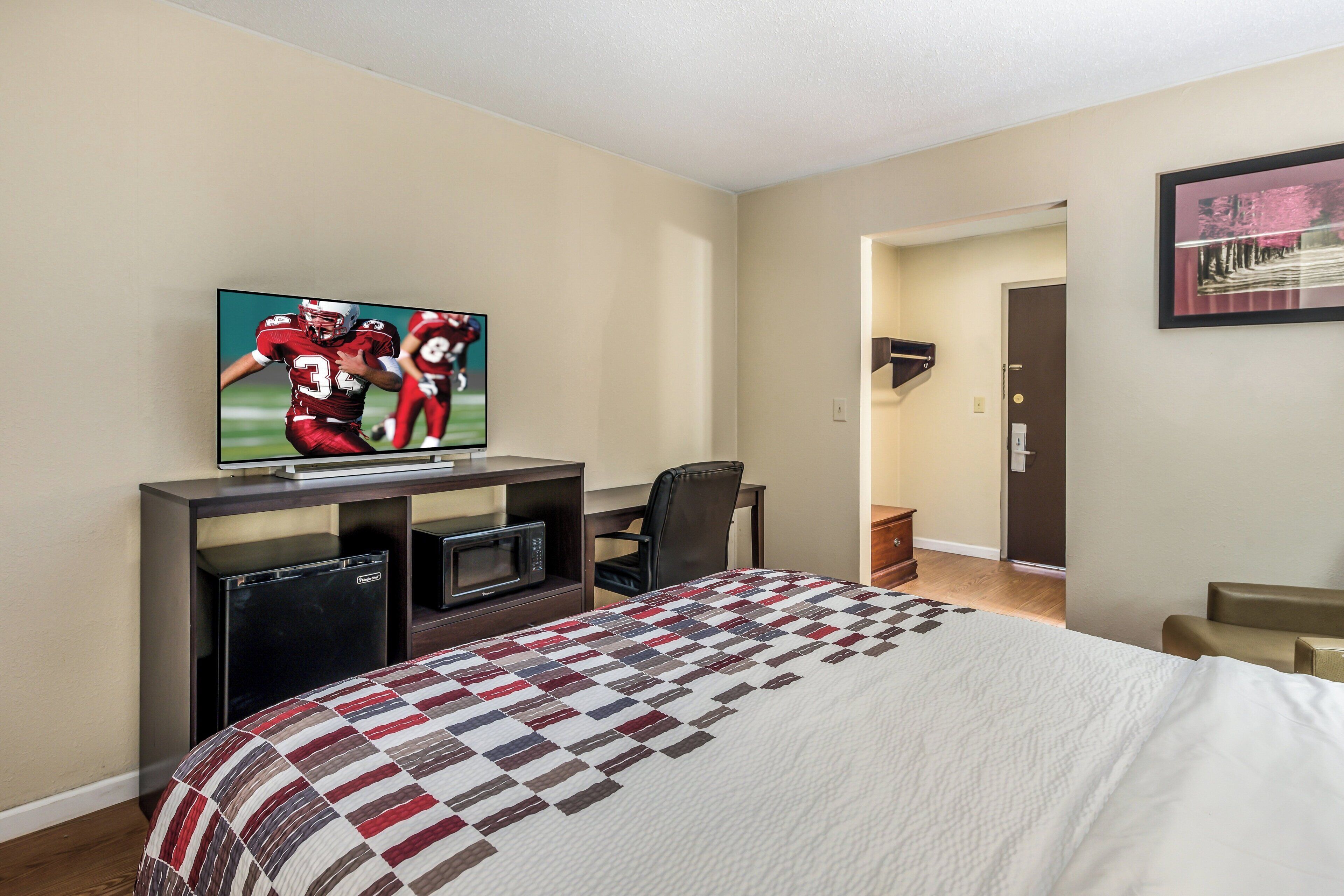 Habitación de hotel con cama, televisor montado en un escritorio, una silla y una puerta que da a otra habitación.