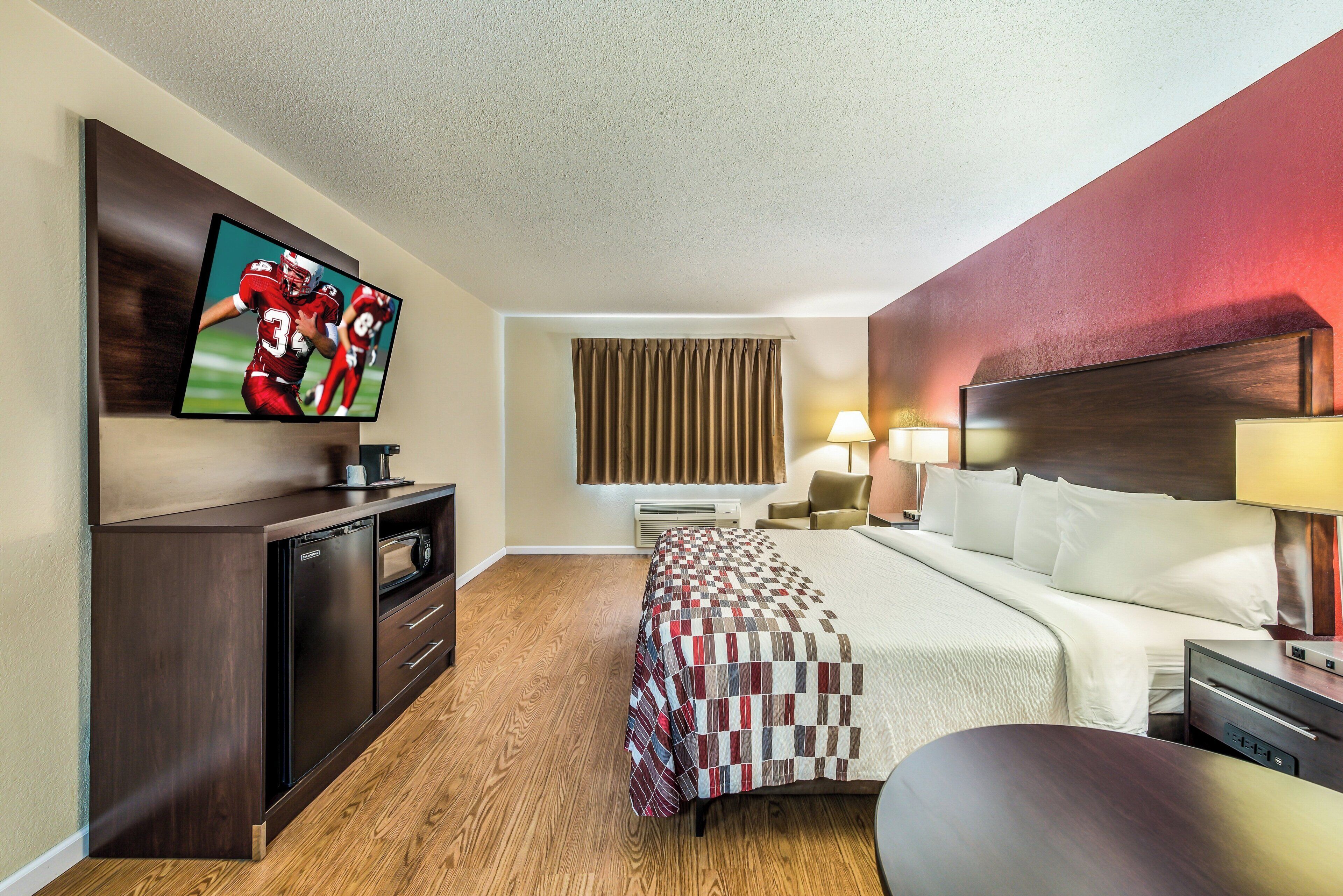 A hotel room with a bed, a desk with a television, a chair, a lamp, and a window with curtains.