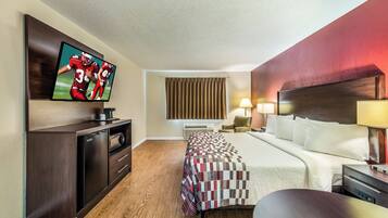 A hotel room with a bed, a desk with a television, a chair, a lamp, and a window with curtains.