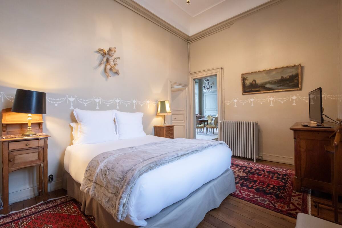 A bedroom with a large bed, two bedside tables with lamps, a radiator, a wooden dresser, and a painting on the wall.