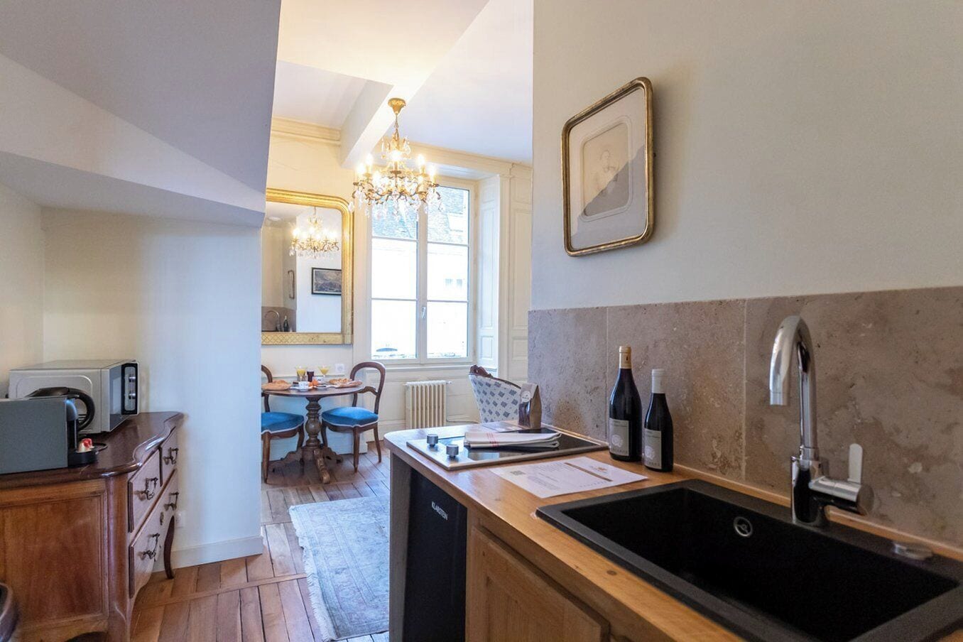 A compact kitchen with a sink, a dining area with a table and chairs, a chandelier, and a framed picture on the wall.