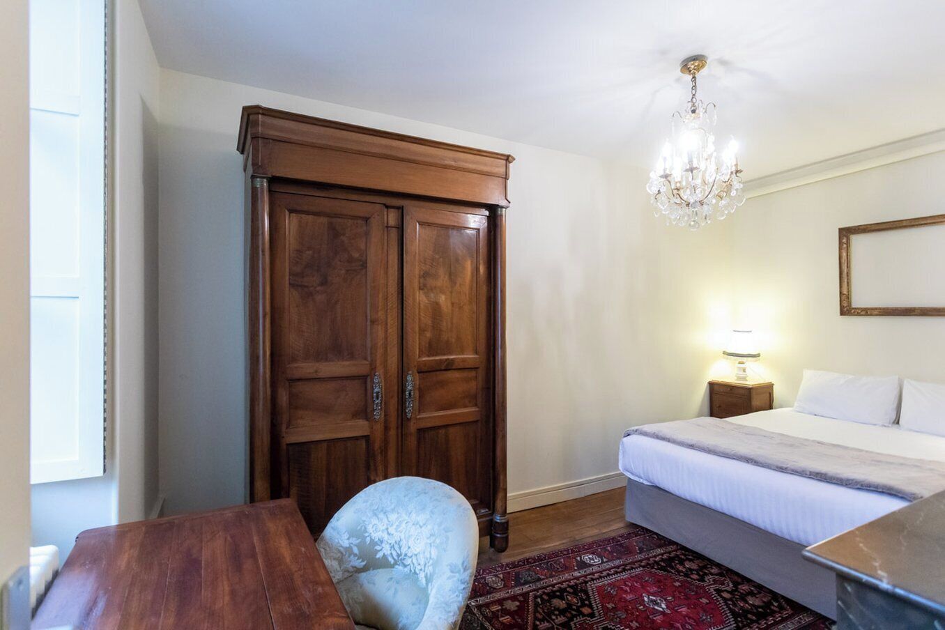 A bedroom with a bed, a wooden wardrobe, a chandelier, a nightstand, and a chair.