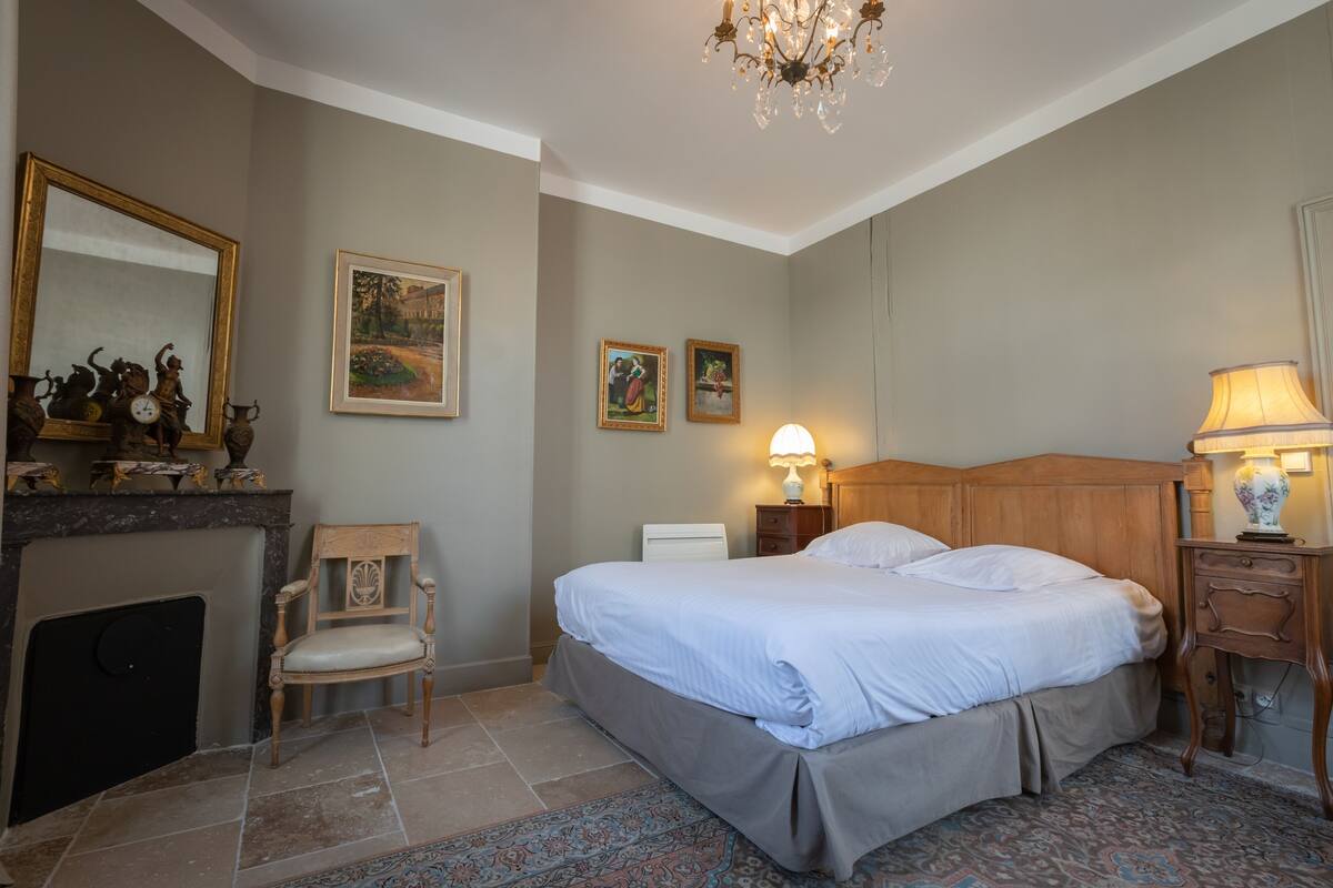 A bedroom with a bed, a chair, a fireplace, and a chandelier.