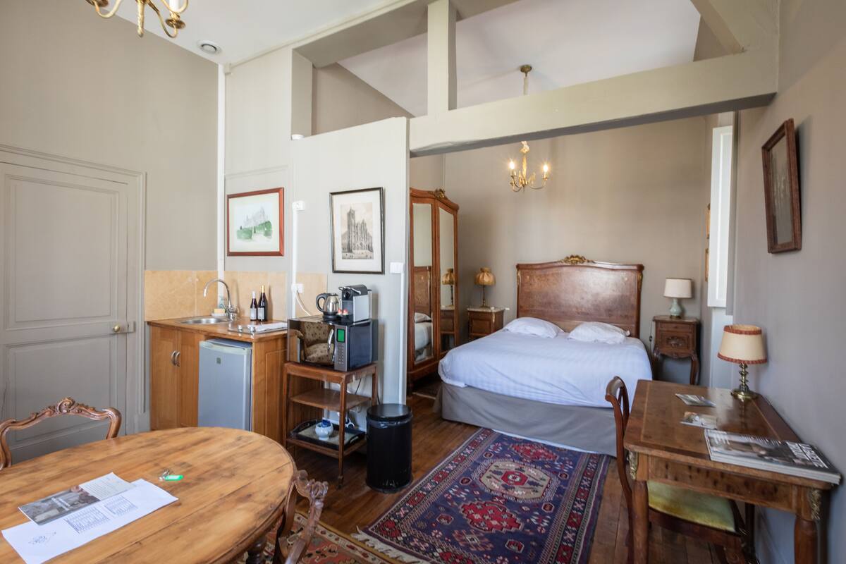 A hotel room with a bed, a desk, a small kitchenette, and a dining table.