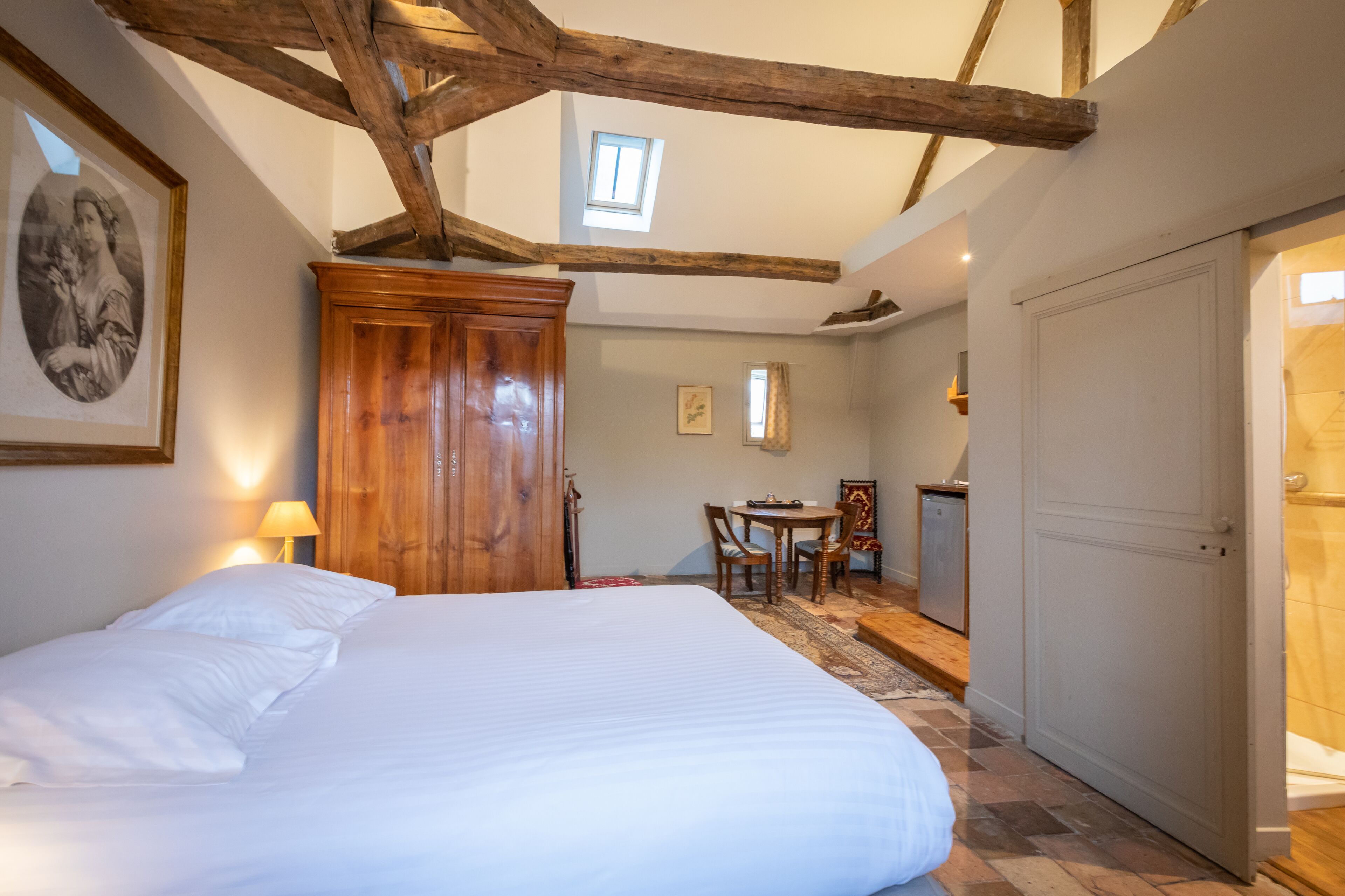 A bedroom with a large bed, wooden ceiling beams, a small table with chairs, and a wardrobe.