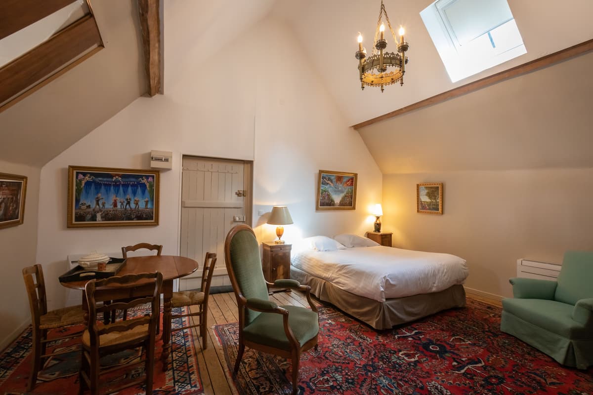 A cozy room with a bed, a dining table with chairs, a chandelier, and artwork on the walls.