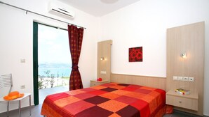 A bedroom with a large bed, a nightstand, a chair, a window with curtains, and a view of the outdoors. - Aptera Paradise (Chania)