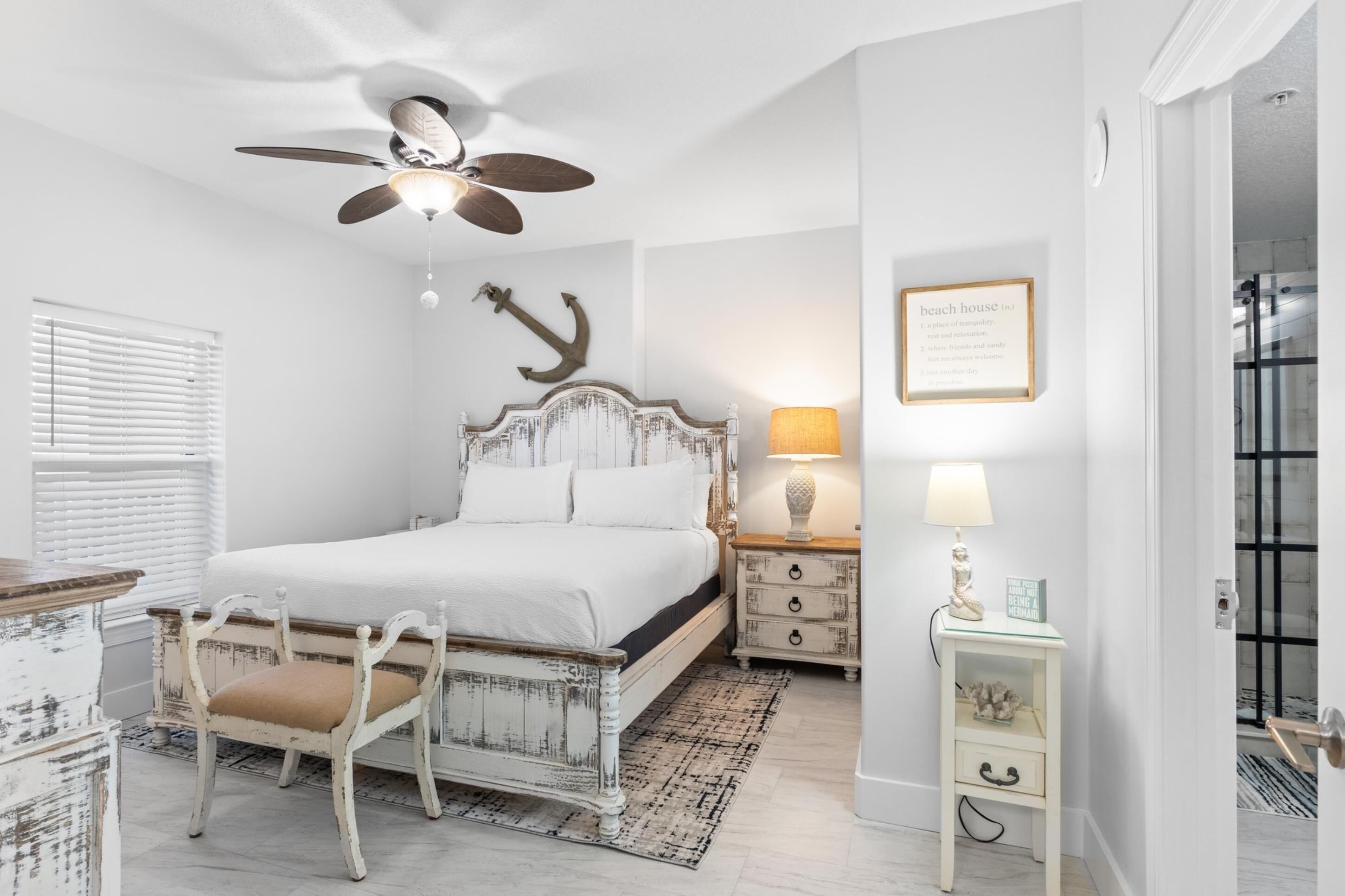 A bedroom with a white bed, a ceiling fan, a nightstand with a lamp, and a framed sign on the wall.