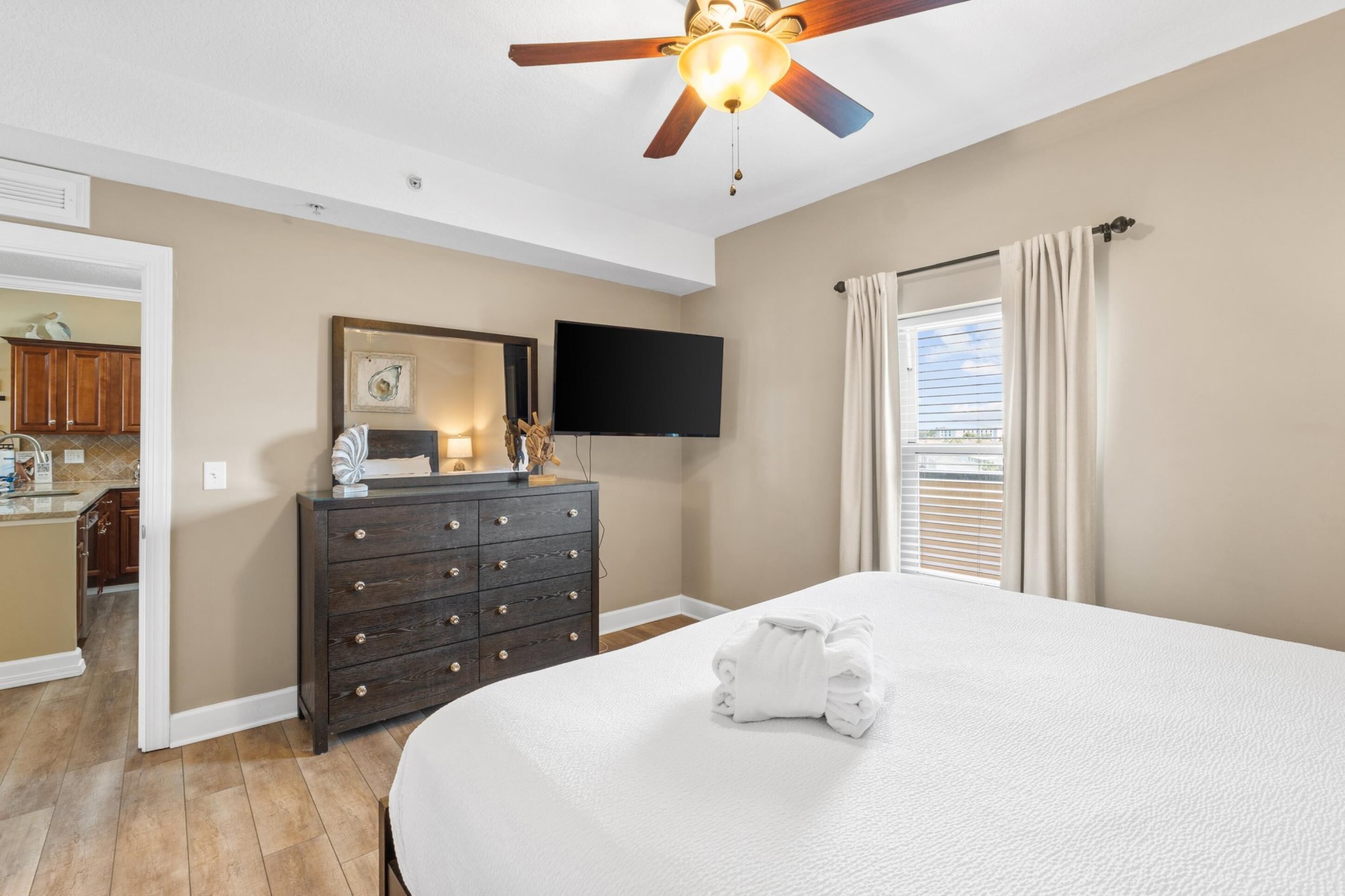A bedroom with a large bed, a ceiling fan, a flat-screen TV, a dresser, and a window with curtains.