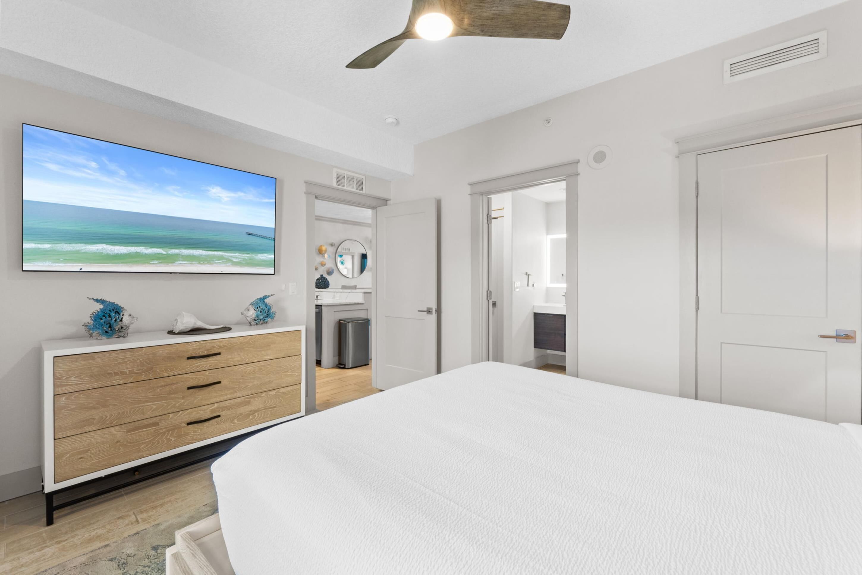 A bedroom with a large bed, a ceiling fan, a TV displaying a beach scene, a wooden dresser, and a view into the bathroom.