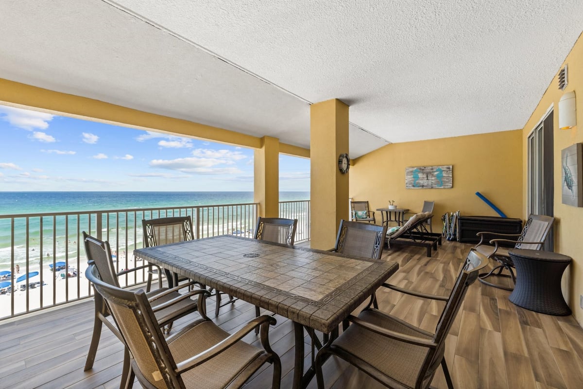 A balcony with a wooden table and chairs, a view of the beach, and a painting on the wall.