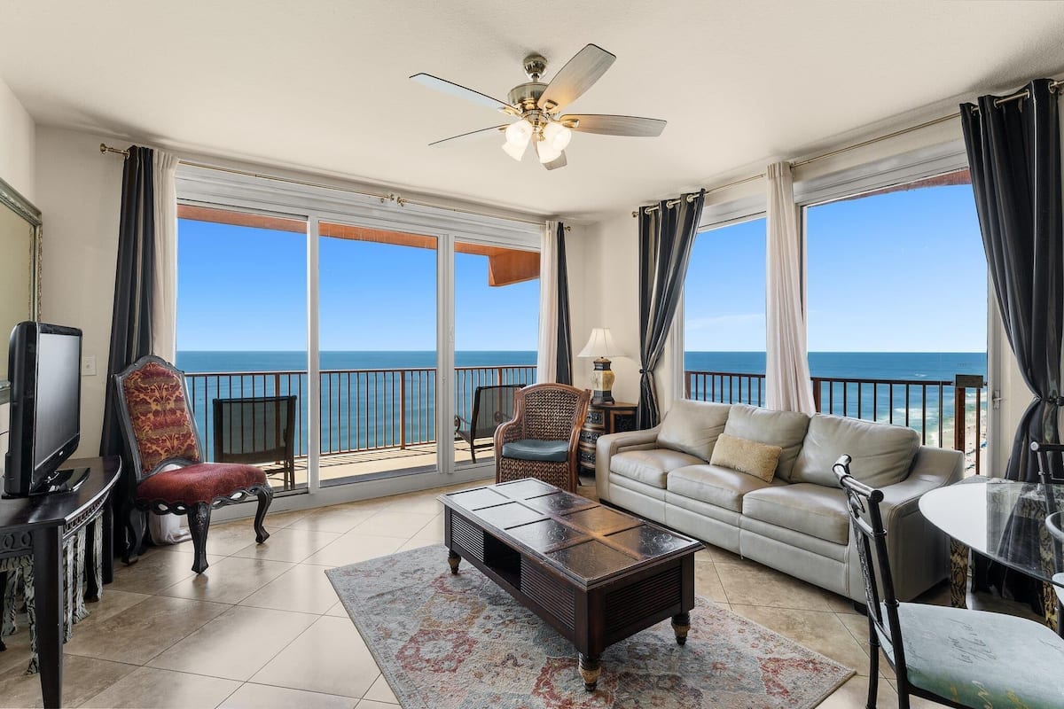 A spacious living room with a large window overlooking the ocean, a ceiling fan, and a television on a stand.