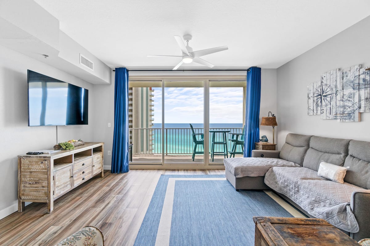 A modern living room with a large flat-screen TV, a ceiling fan, a sofa, a coffee table, and a view of the ocean through a sliding glass door.