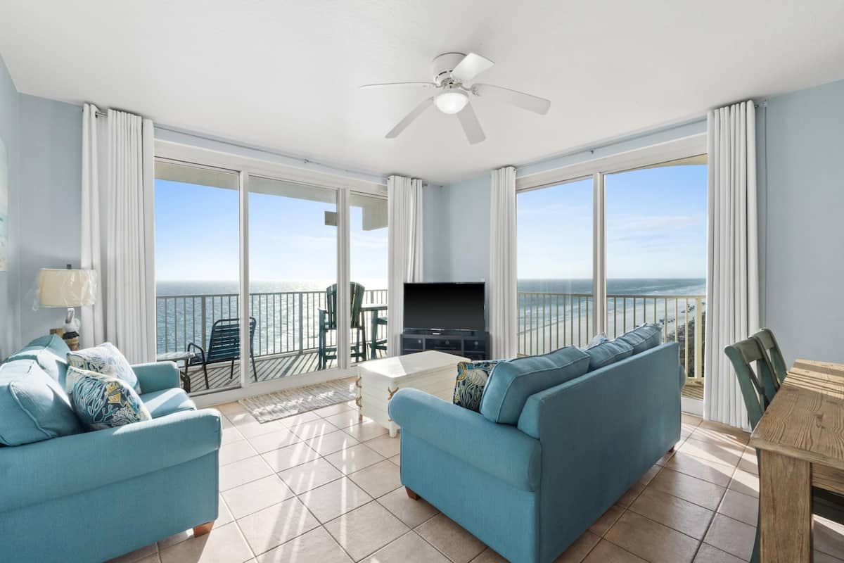 A spacious living room with a large window overlooking the sea, a ceiling fan, and a dining area with a table and chairs.