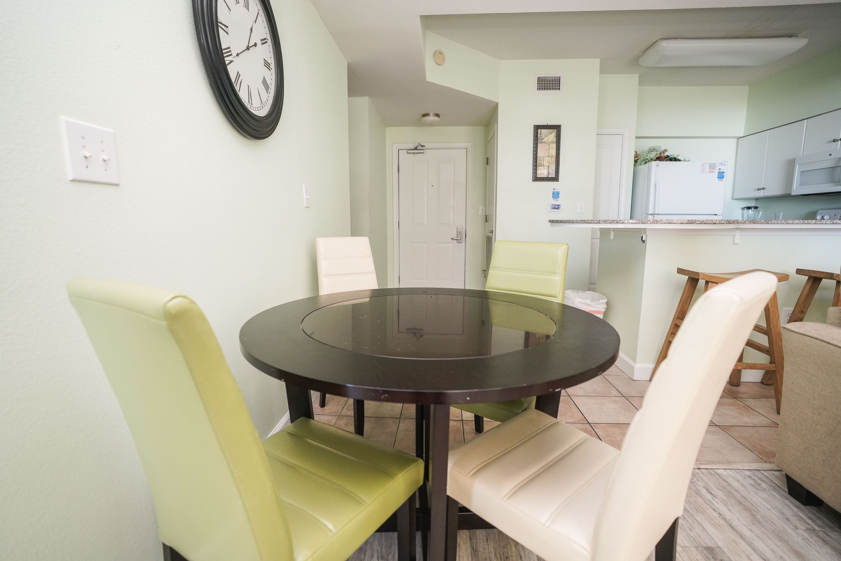 A dining area with a round table and four chairs, a kitchen in the background, and a clock on the wall.
