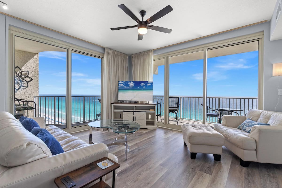 A spacious living room with a large TV, comfortable seating, a glass coffee table, and a ceiling fan.