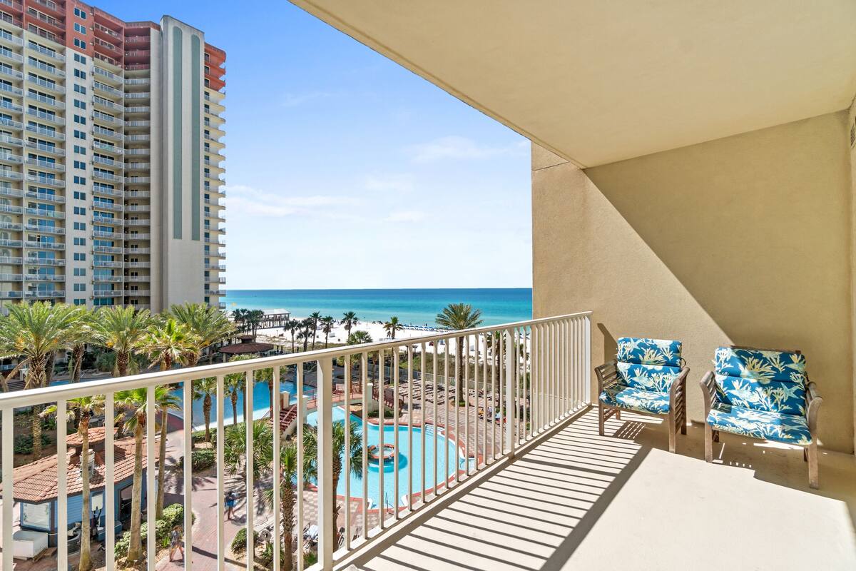 A balcony with a pool, beach view, and two chairs.