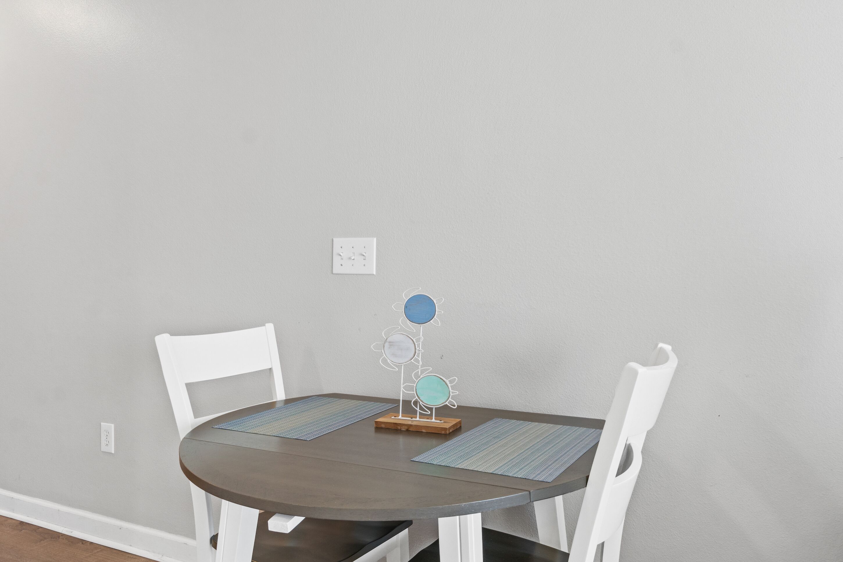 A round dining table with two white chairs, a decorative metal flower stand, and two placemats.