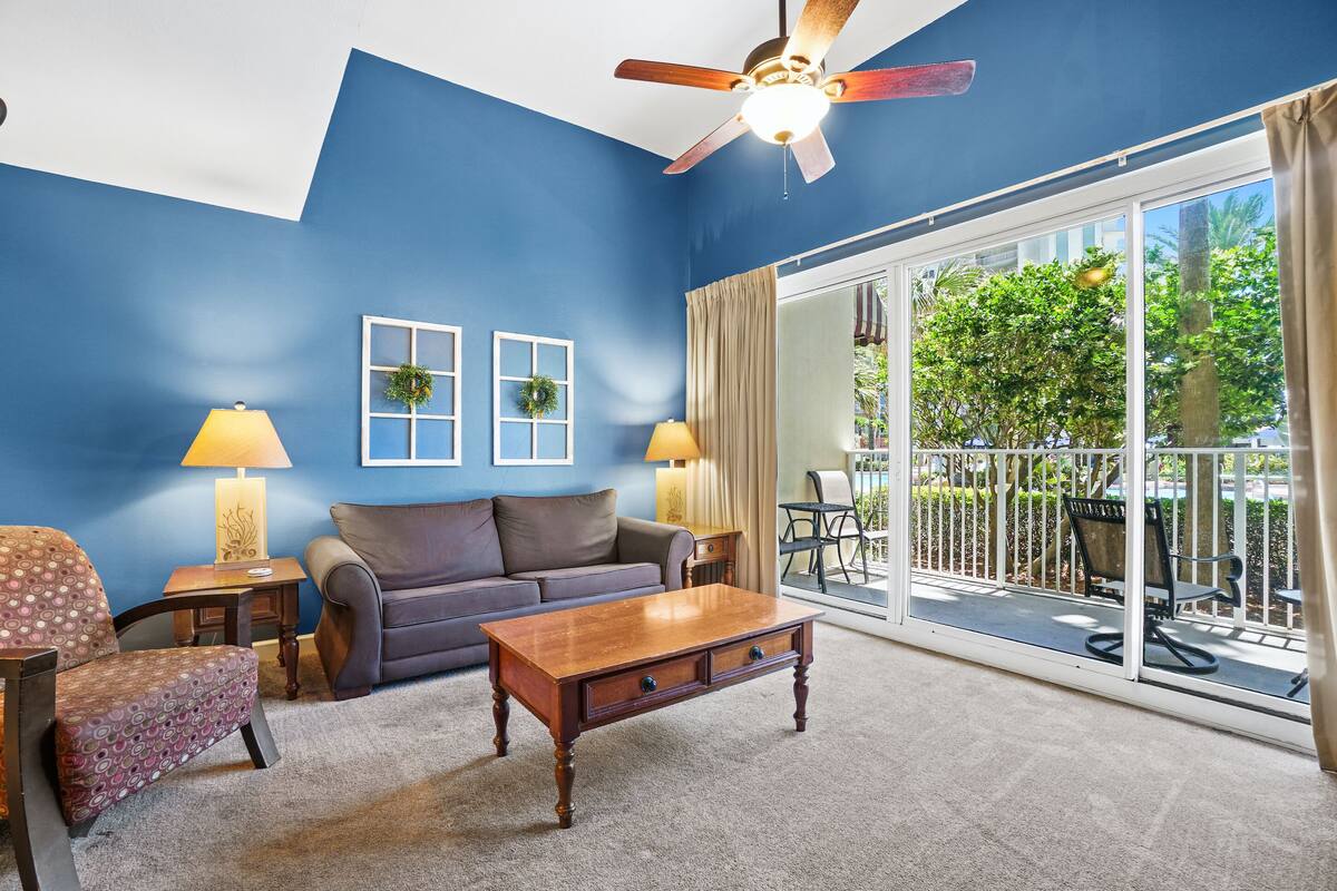 A living room with a blue wall, a ceiling fan, a sofa, a coffee table, and a view of a balcony with outdoor furniture.