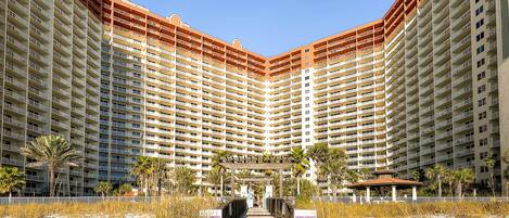 Un hotel grande con varios pisos, un paseo central y una zona de playa con palmeras.