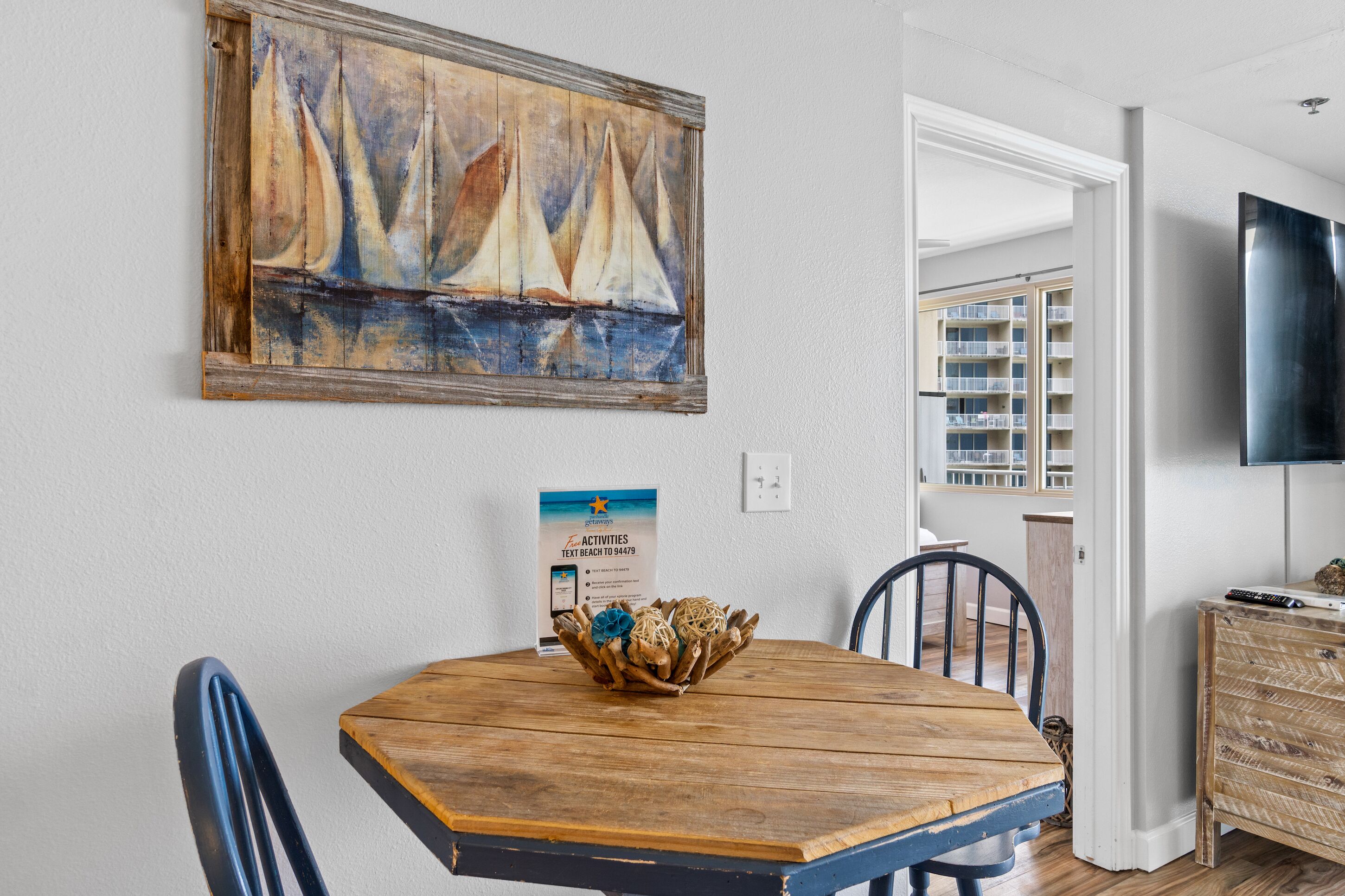 A dining area with a wooden table, chairs, a painting, a sign, and a view of a balcony with a TV.