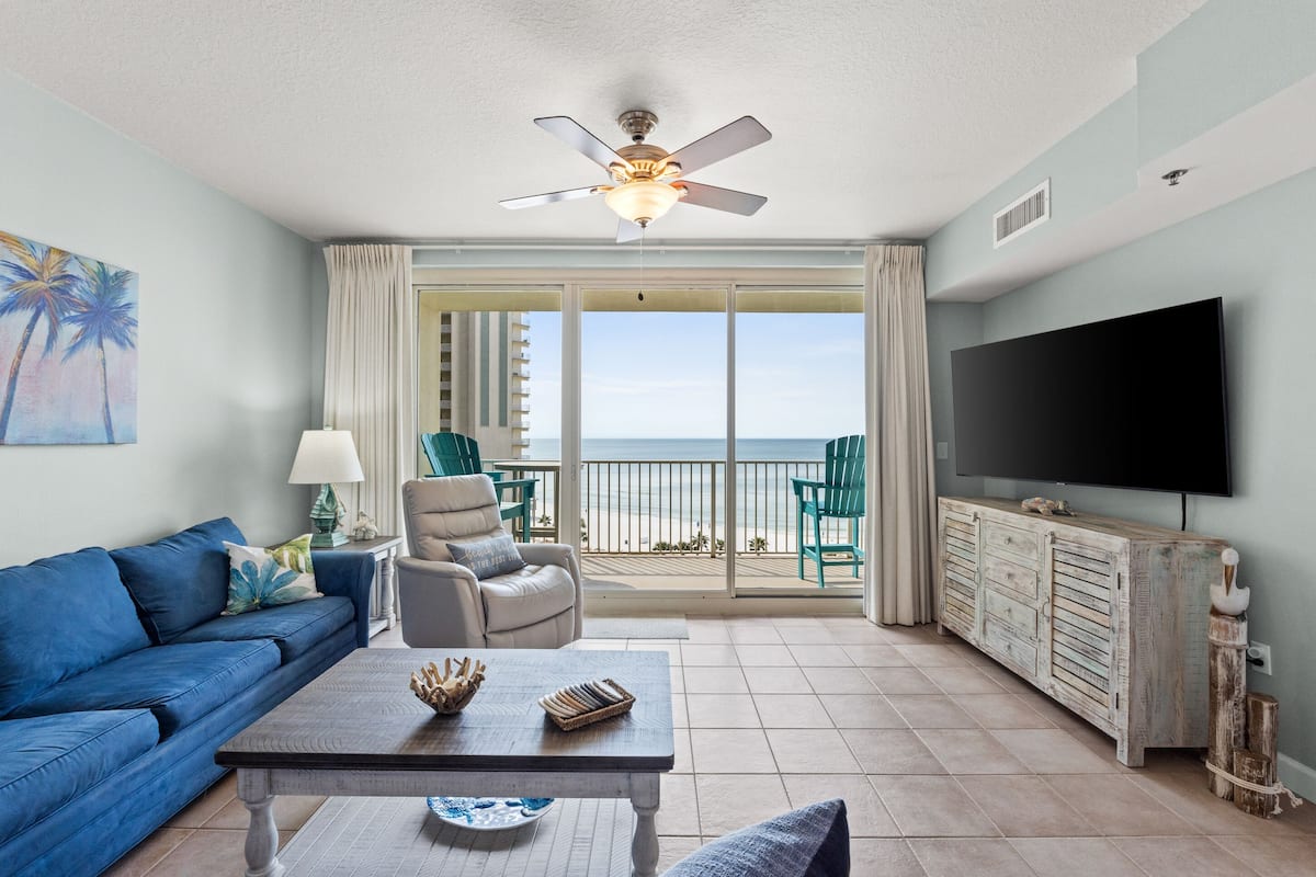 A living room with a blue sofa, a coffee table, a ceiling fan, a TV, and a balcony with a view of the ocean.