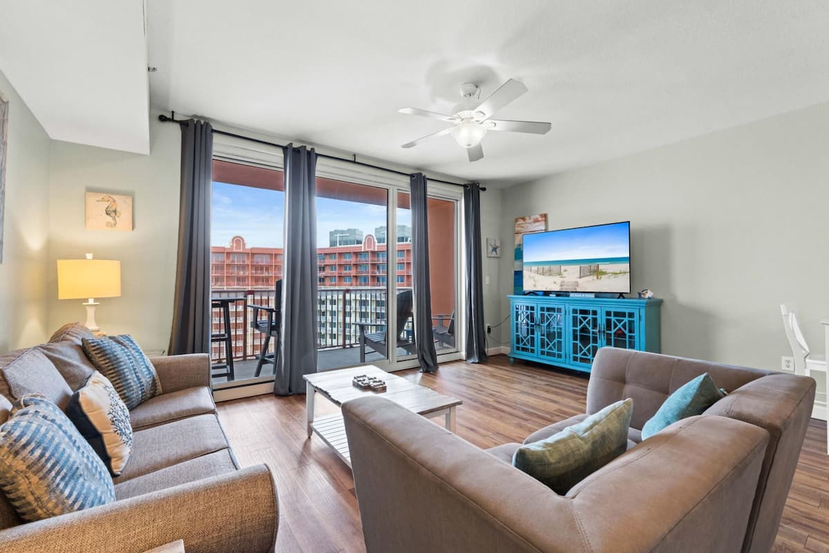 A living room with a sofa, a coffee table, a TV, and a balcony with a view of buildings.
