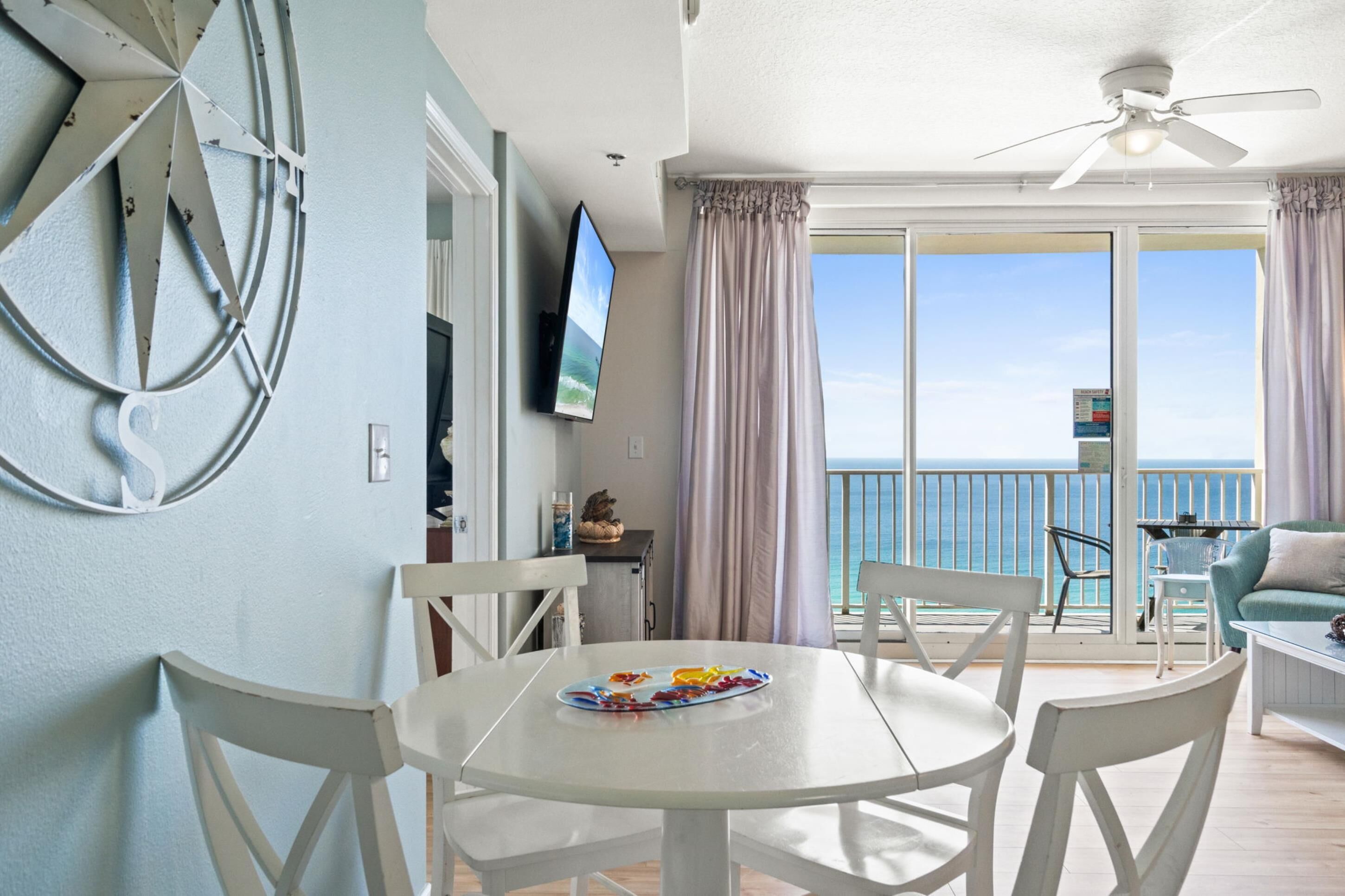 A room with a round white table, white chairs, a flat-screen TV, and a balcony with a view of the sea.