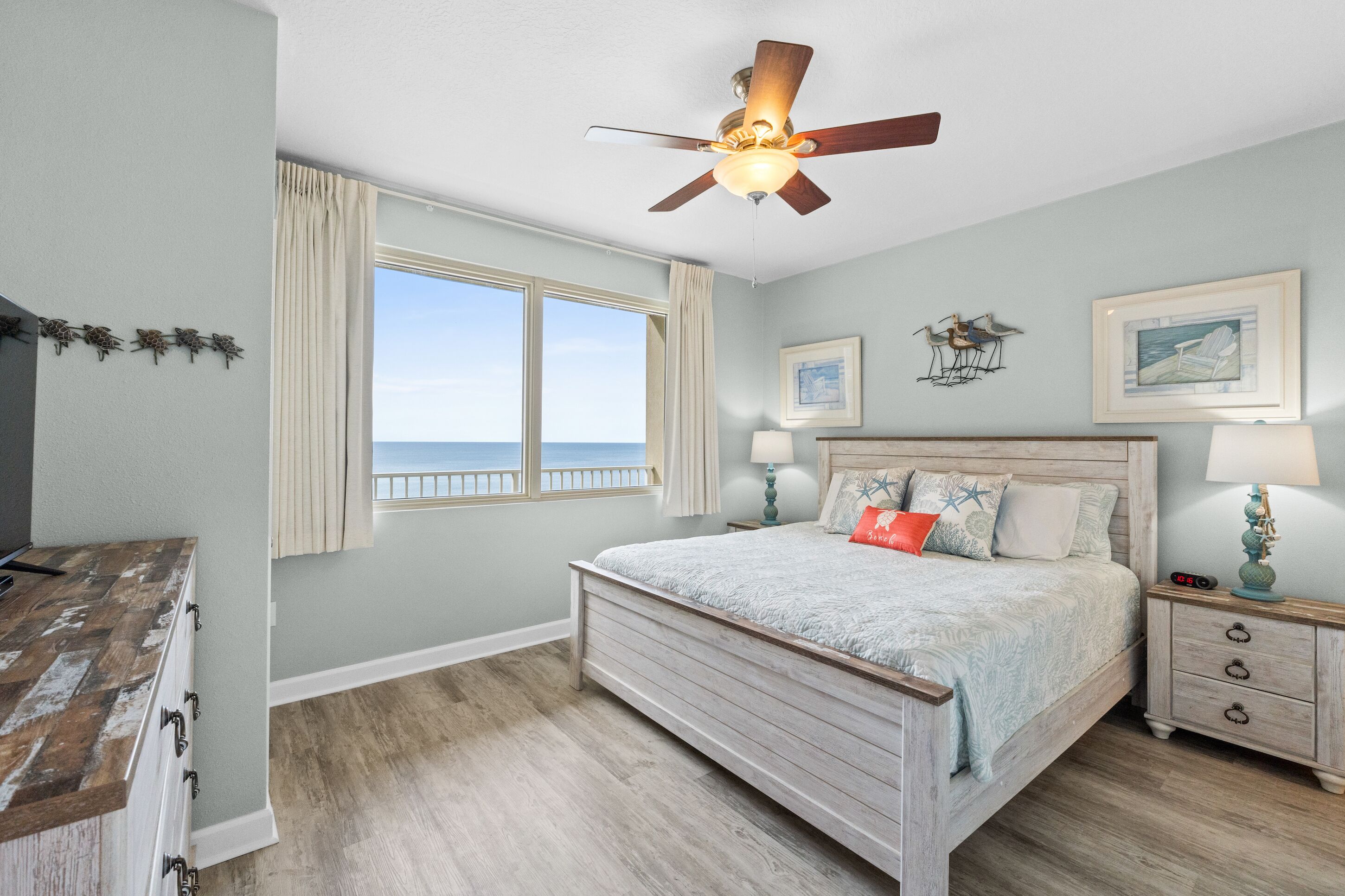 A bedroom with a large bed, a ceiling fan, a nightstand, and a window with a view of the ocean.