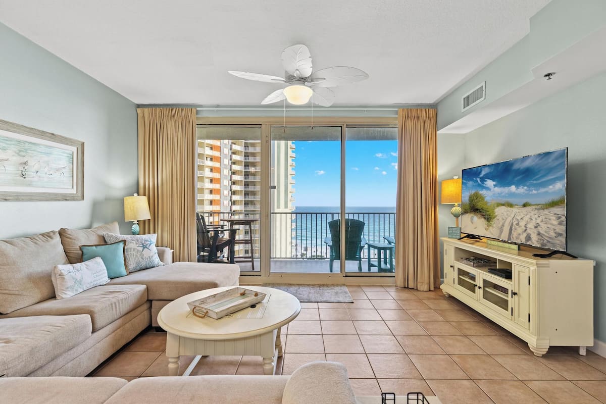 A living room with a sofa, coffee table, TV, and balcony with a view of the ocean.
