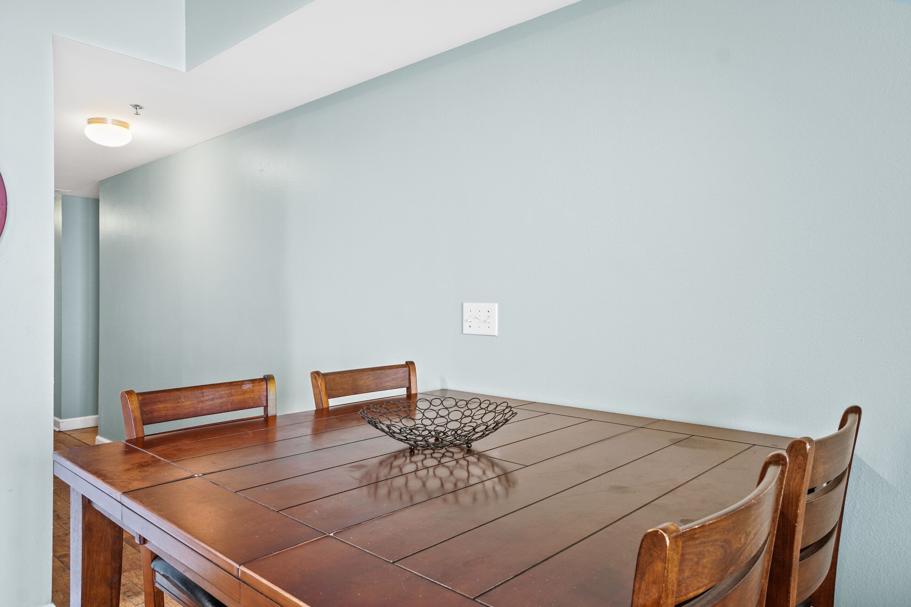 Una mesa de comedor con cuatro sillas de madera, un cuenco metálico decorativo y una pared azul claro con una lámpara de techo.