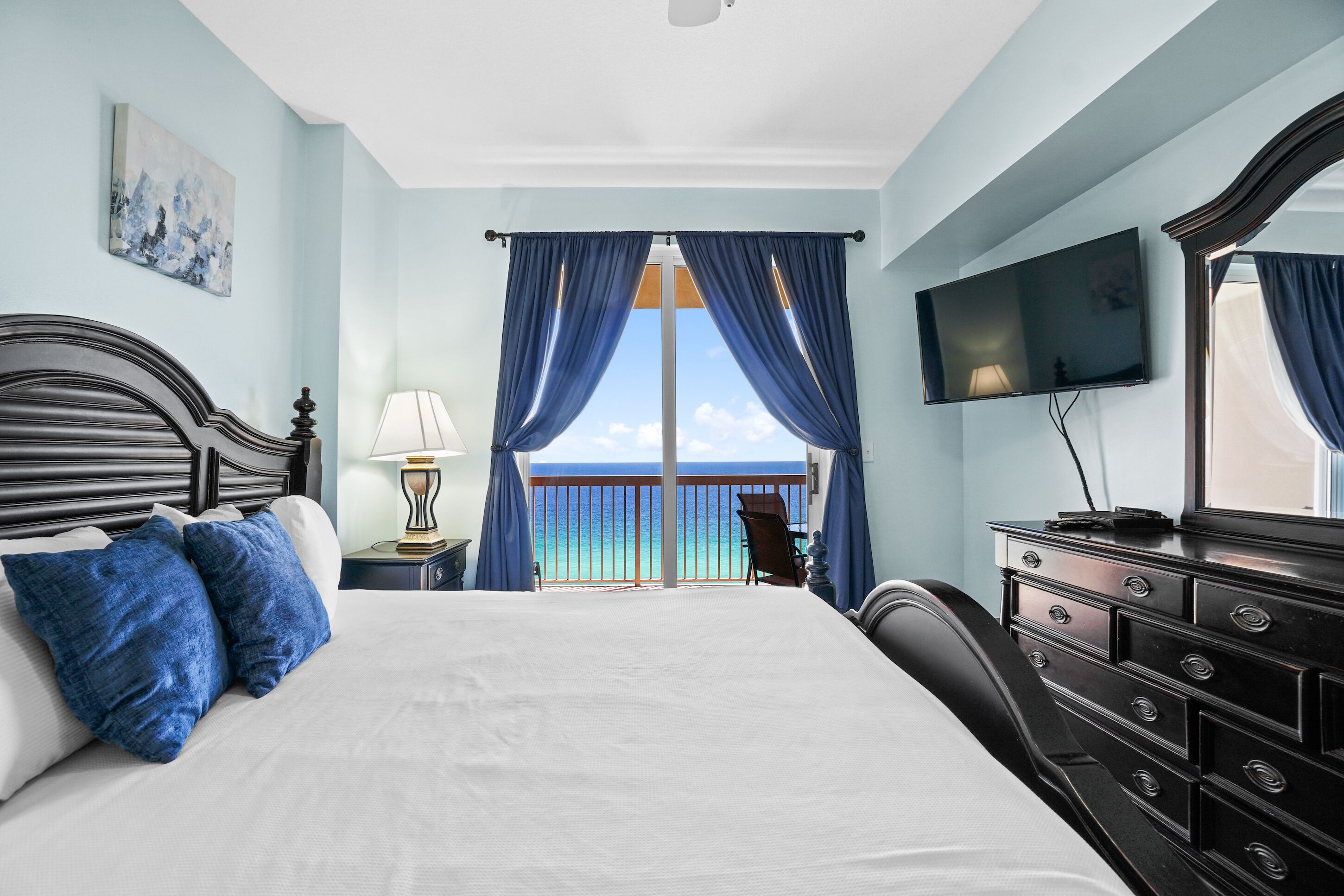 A bedroom with a large bed, a dresser, a television, and a balcony with a view of the ocean.