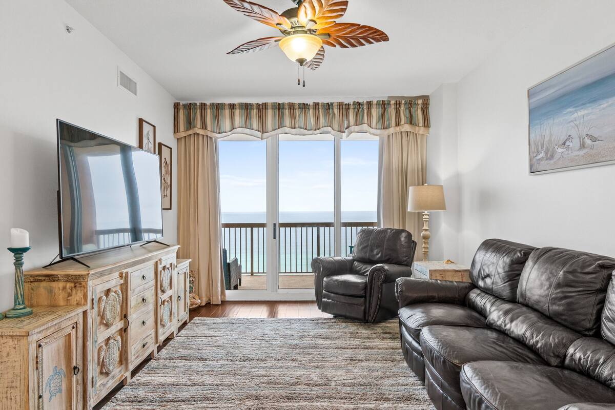 A living room with a large flat-screen TV, a leather sofa, a ceiling fan, and a view of the ocean through large windows.