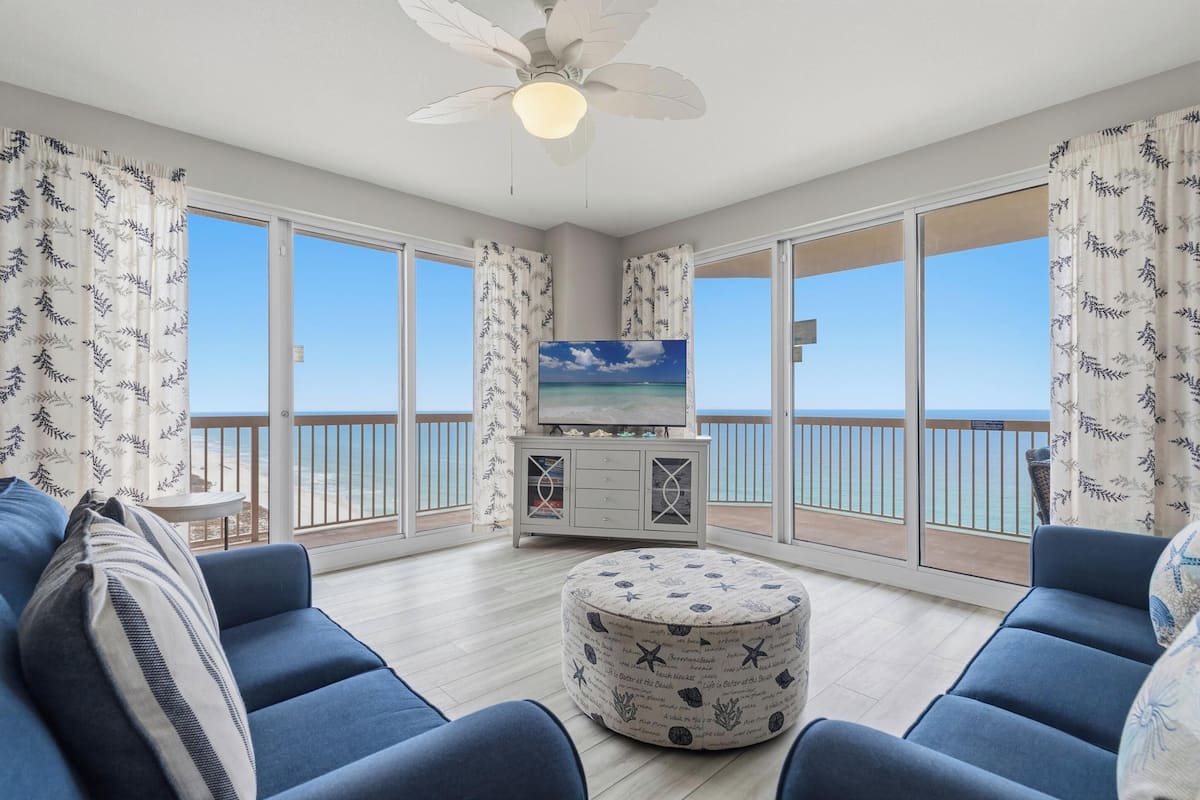 A balcony with a TV, blue sofas, a round ottoman, and a ceiling fan.