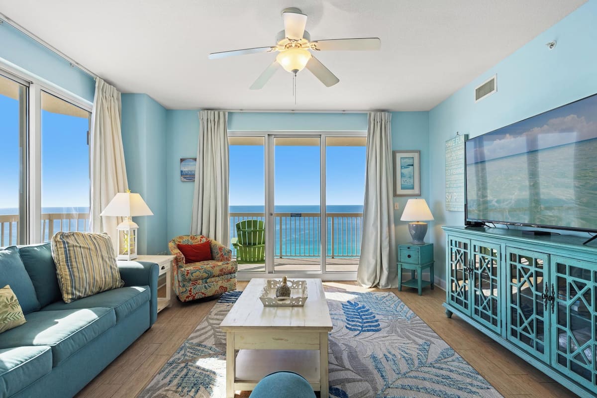 A living room with a sofa, a coffee table, a television, and a view of the ocean through large windows.