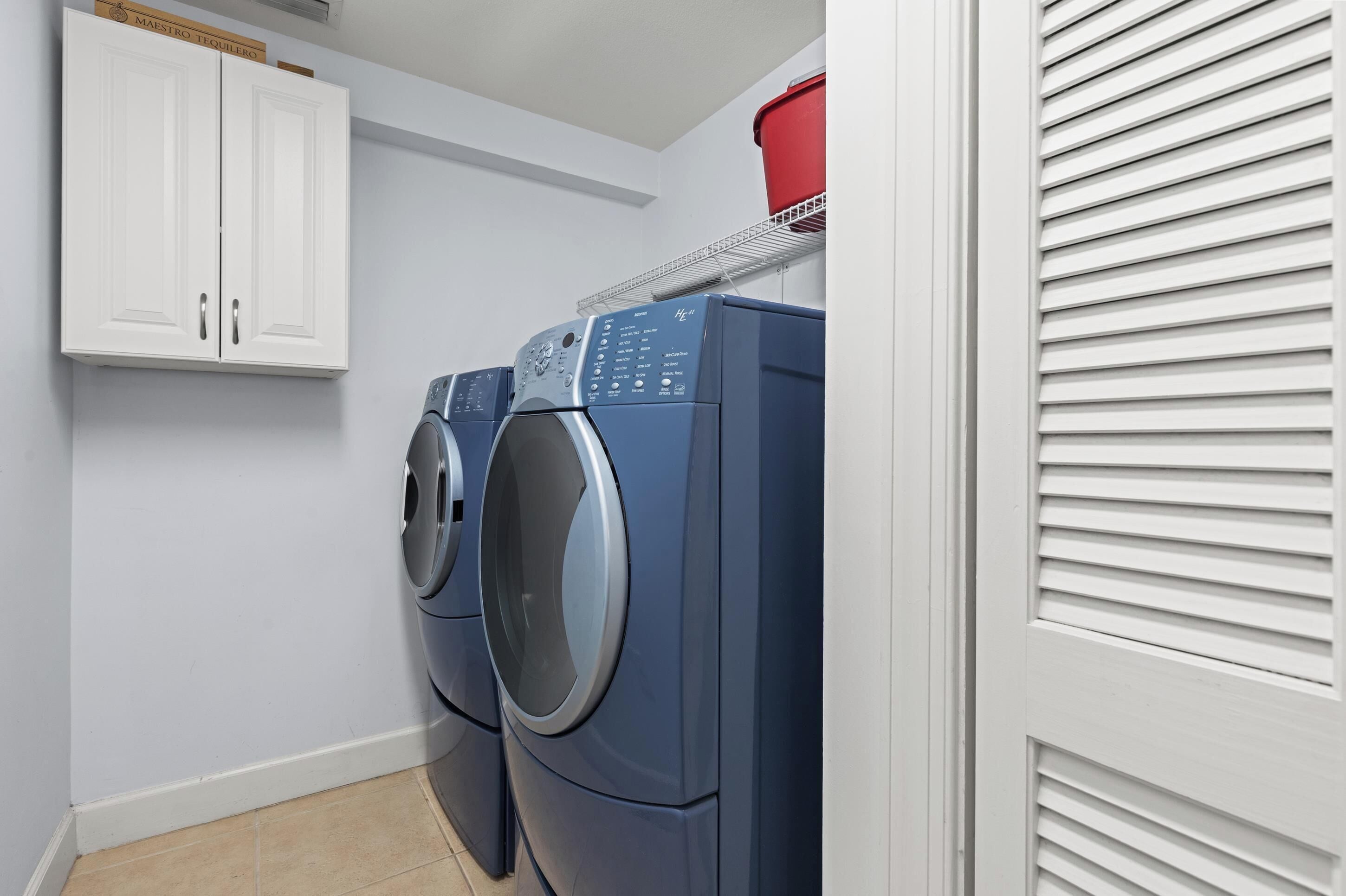 A laundry room with two front-loading washing machines, a white cabinet, and a red container on a shelf.
