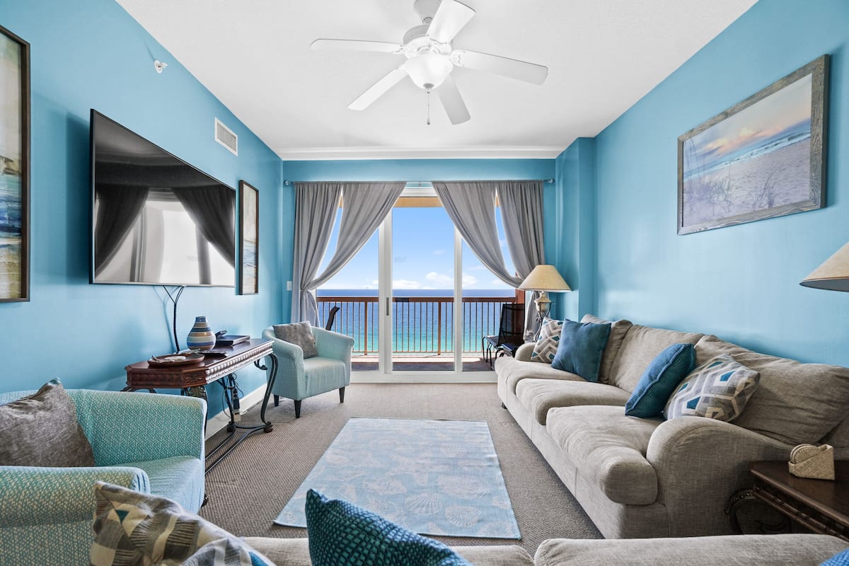 A living room with a large window, a ceiling fan, and a view of the ocean.