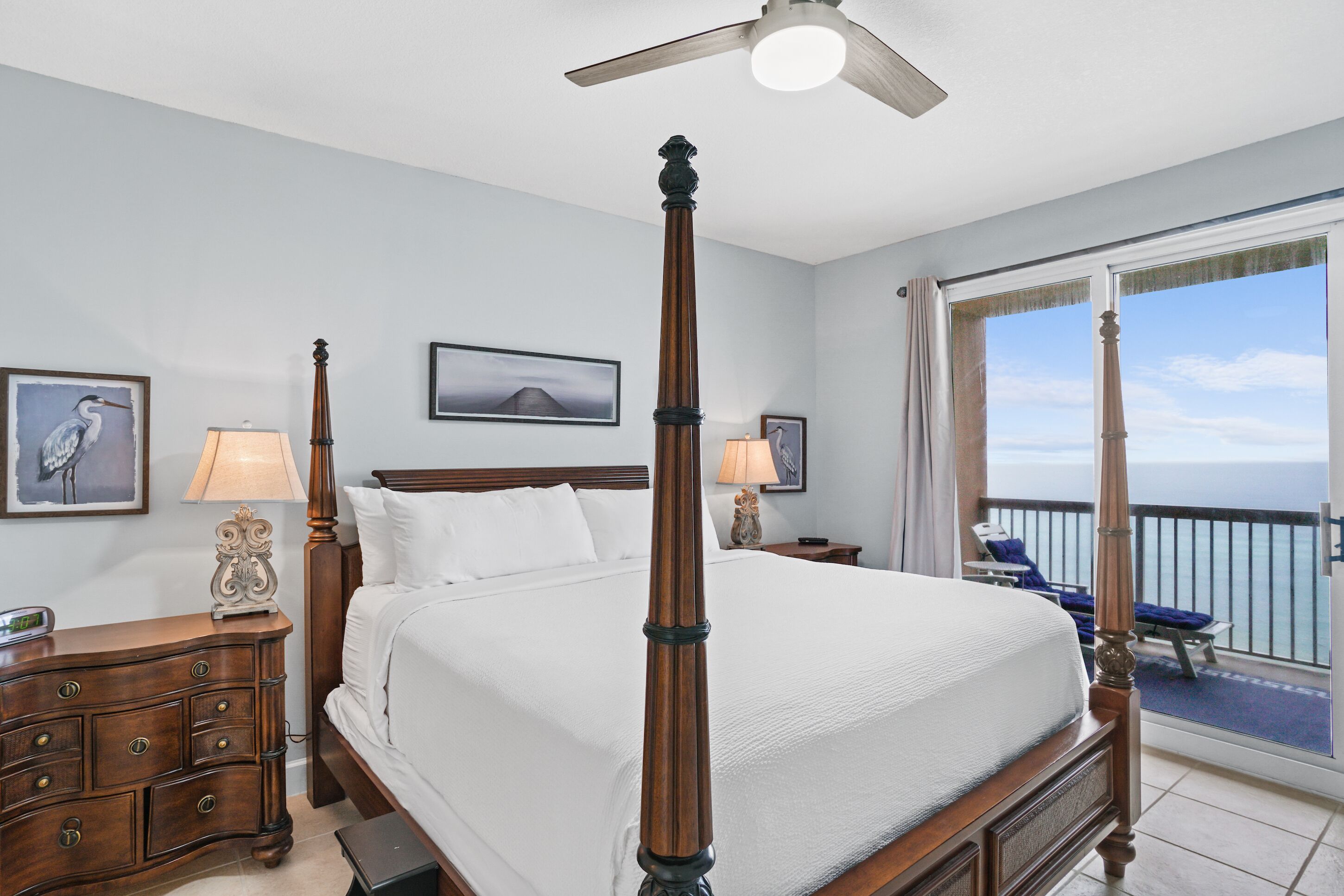 A four-poster bed with a ceiling fan, a nightstand with a lamp, and a view of the ocean through a balcony.