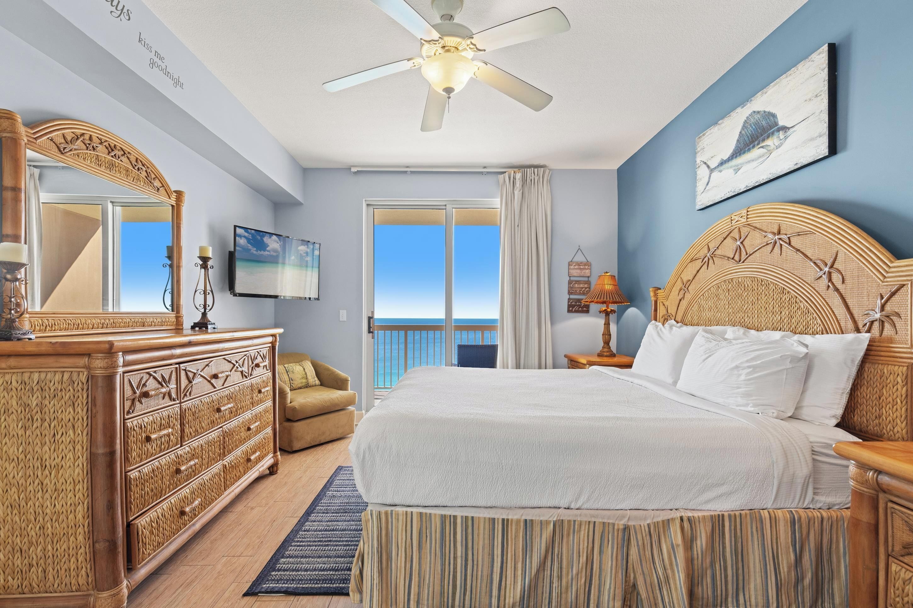 A bedroom with a large bed, a ceiling fan, a wooden dresser, a nightstand, and a view of the ocean.