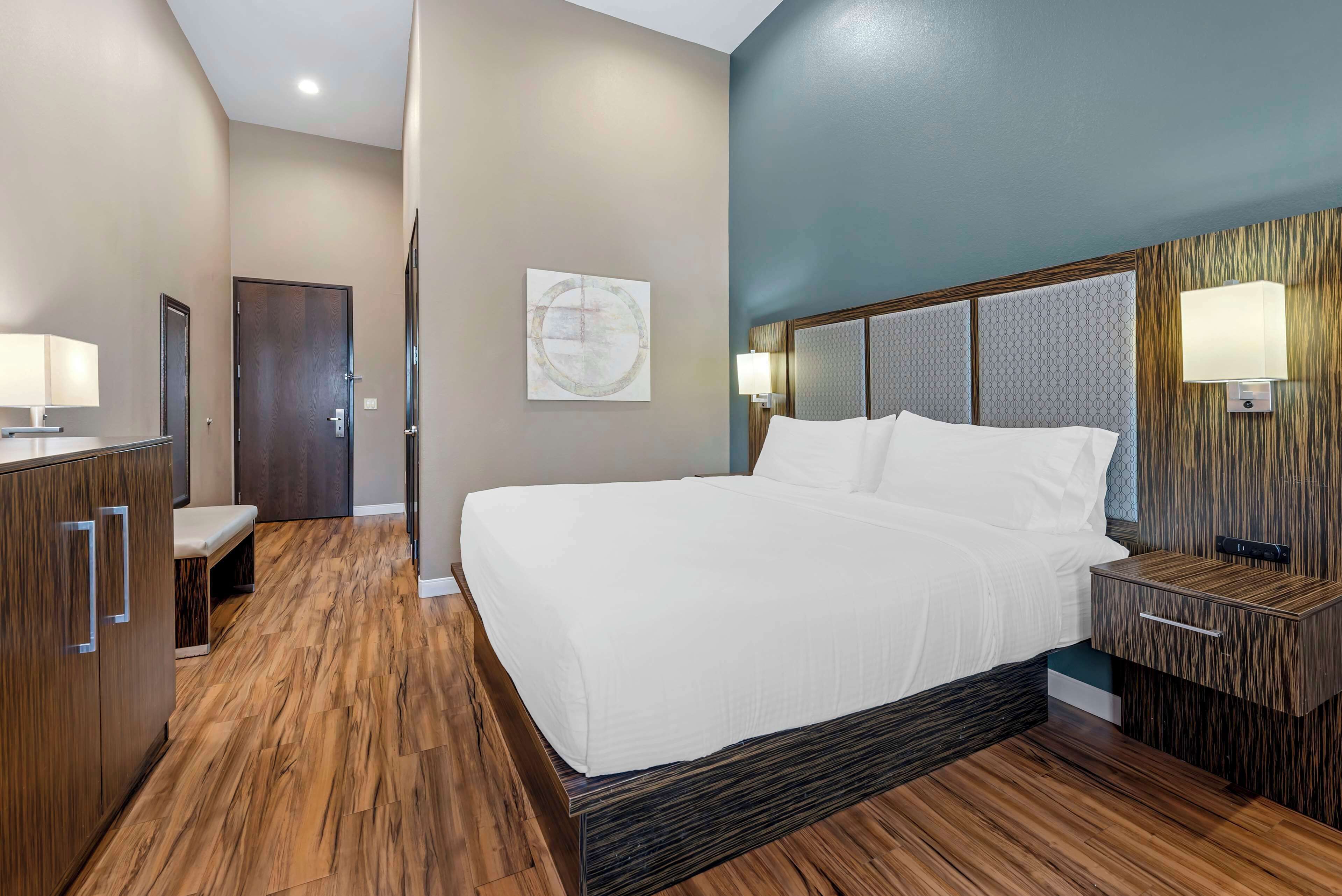 Quarto de hotel com uma cama grande, piso de madeira, uma escrivaninha com abajur e luminária na parede.
