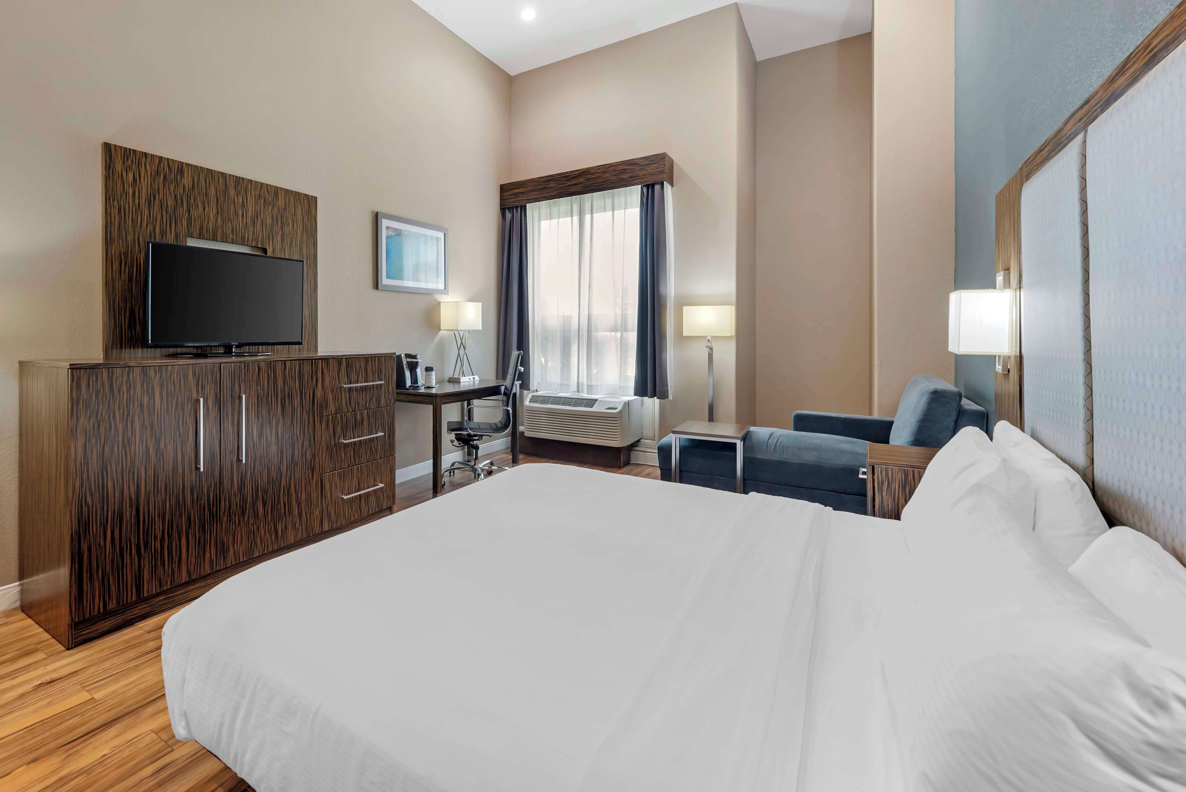 Habitación de hotel con una cama grande, un televisor sobre un mueble de madera, un escritorio con una silla, una ventana con cortinas y una mesita de noche con lámpara.