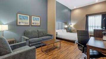 Une chambre d’hôtel avec un canapé gris, une table basse en bois, un lit avec du linge de lit blanc, un fauteuil de bureau noir et un bureau avec une lampe.