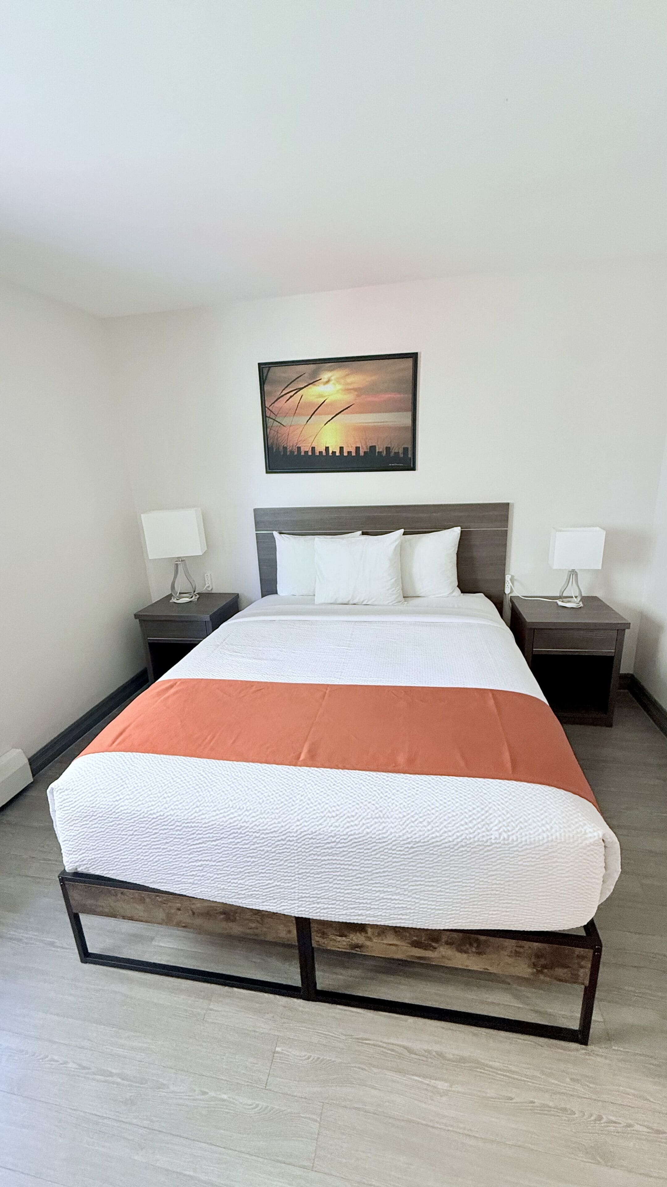 Una habitación de hotel con una cama grande, dos mesitas de noche, un buró, un cuadro en la pared y un suelo de color claro.
