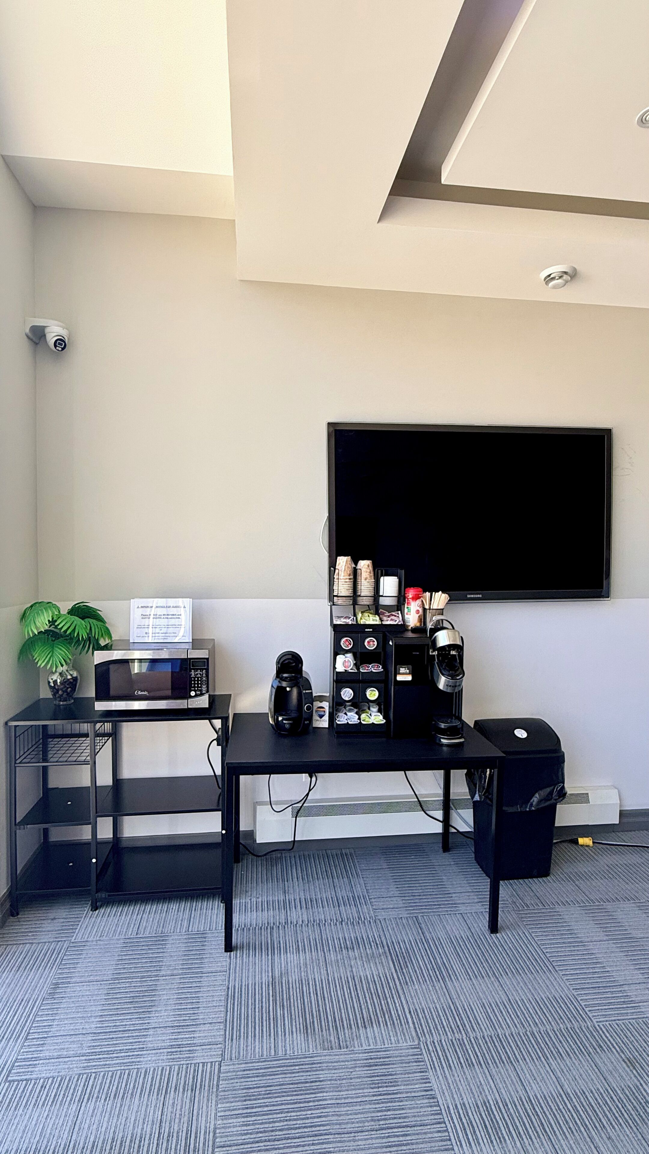 Una pequeña oficina con un escritorio, microondas, máquina de café, cubo de basura y televisor montado en la pared.
