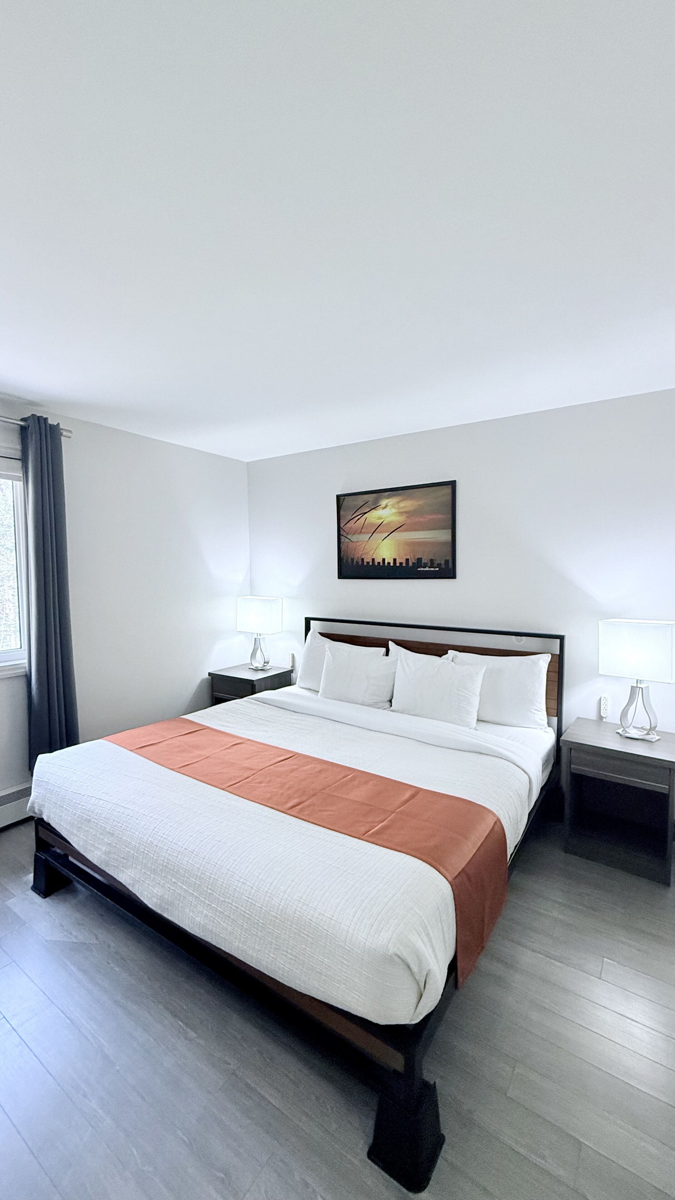 A hotel room with a large bed, two bedside tables, a nightstand, a window with curtains, and a cityscape painting on the wall.