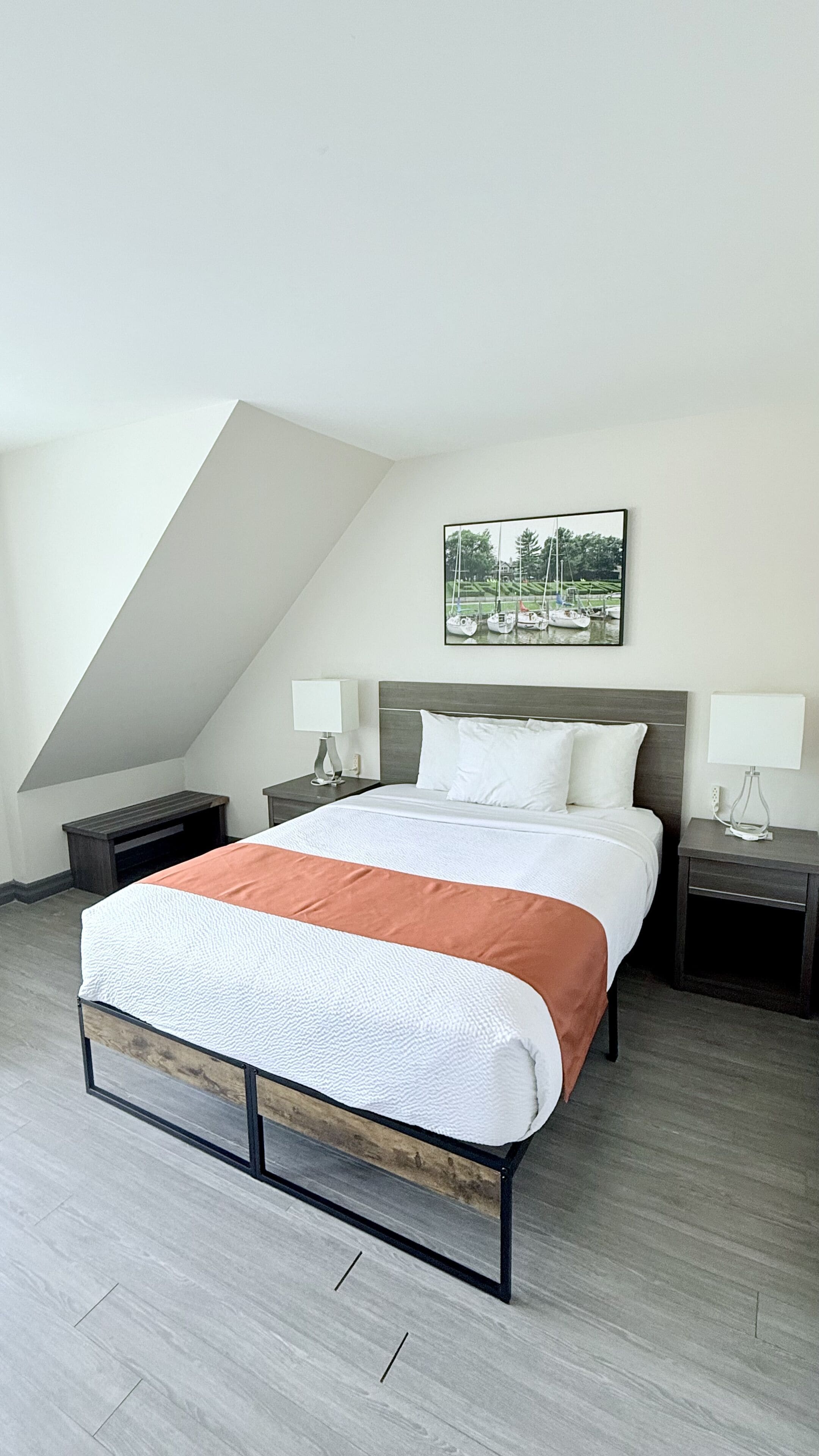 A modern bedroom with a sloped ceiling, a large bed with a white and orange bedspread, two bedside tables with lamps, and a framed picture on the wall.